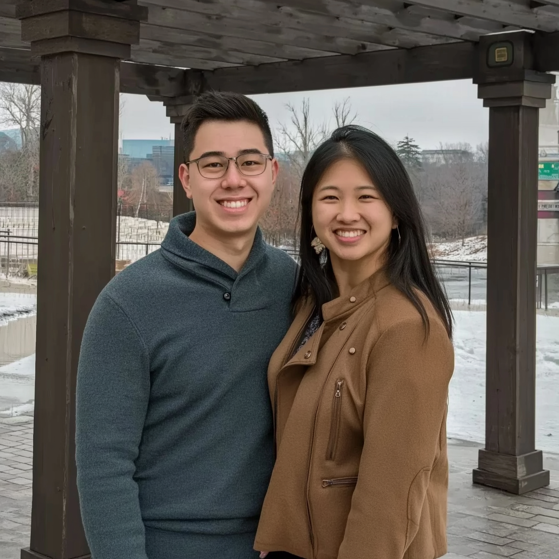 Smiling couple standing near wooden structure outdoors. Man wears glasses and a blue sweater, woman wears a brown jacket.