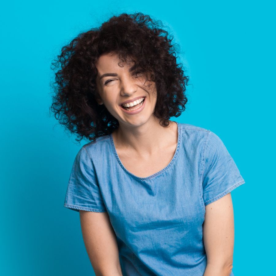 A woman with curly hair is laughing and wearing a blue shirt.