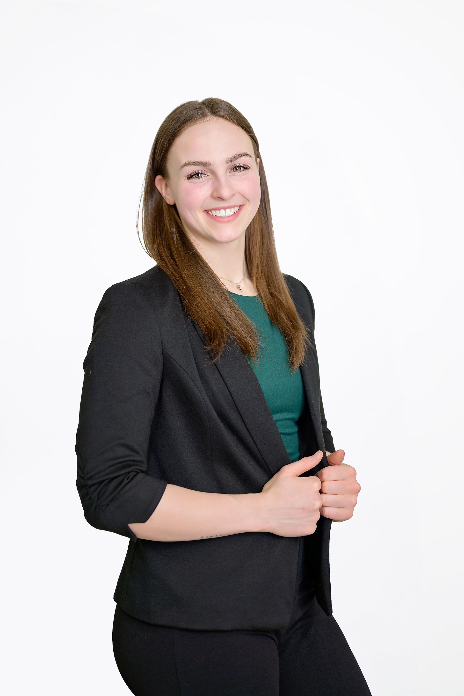 Femme en blazer noir souriante, tenant sa veste, haut vert, fond blanc.