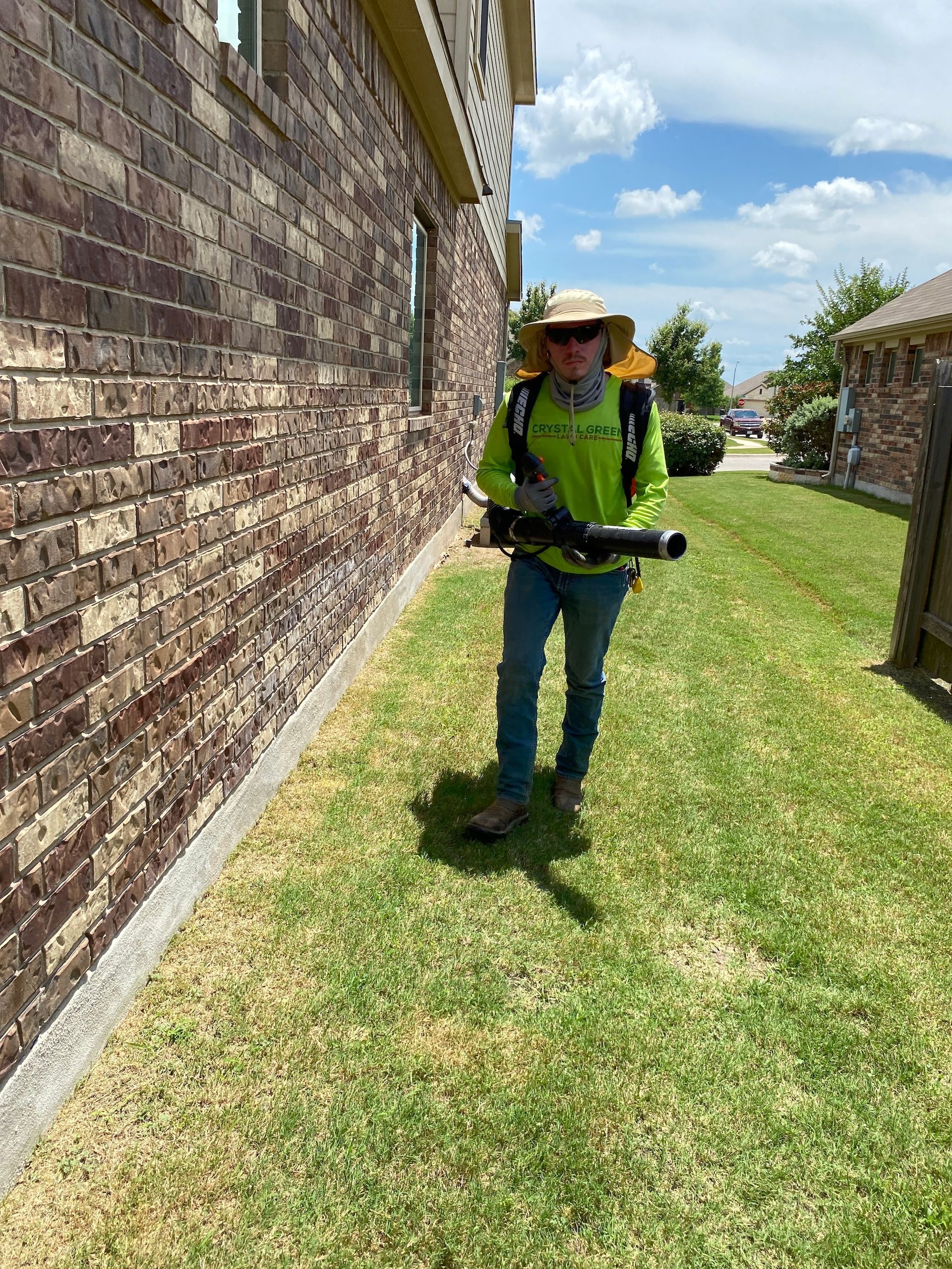 A man is standing in the grass holding a blower in front of a brick building.