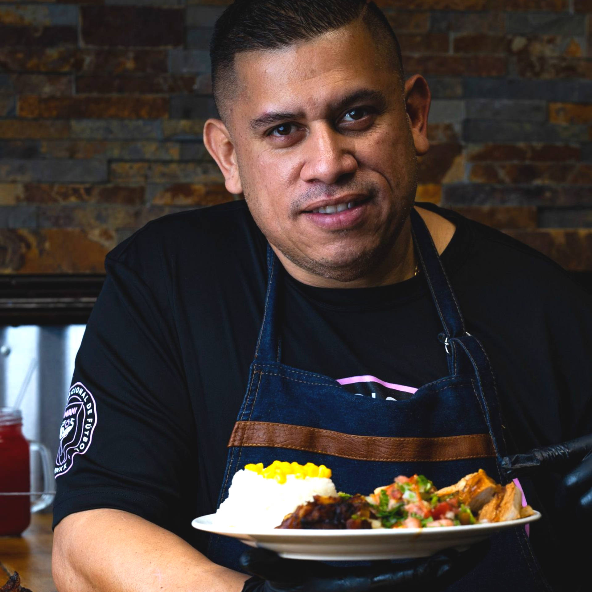 Chef holding plate with food; wearing apron, smiling at the camera. Plate has rice, vegetables, and meat.