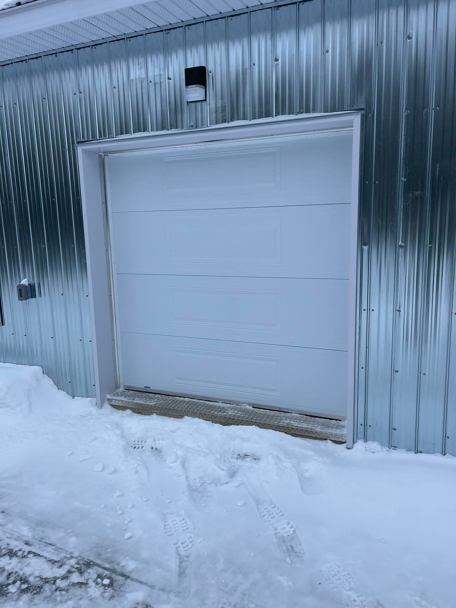 Une porte de garage blanche est ouverte dans la neige à côté d'un bâtiment en métal.