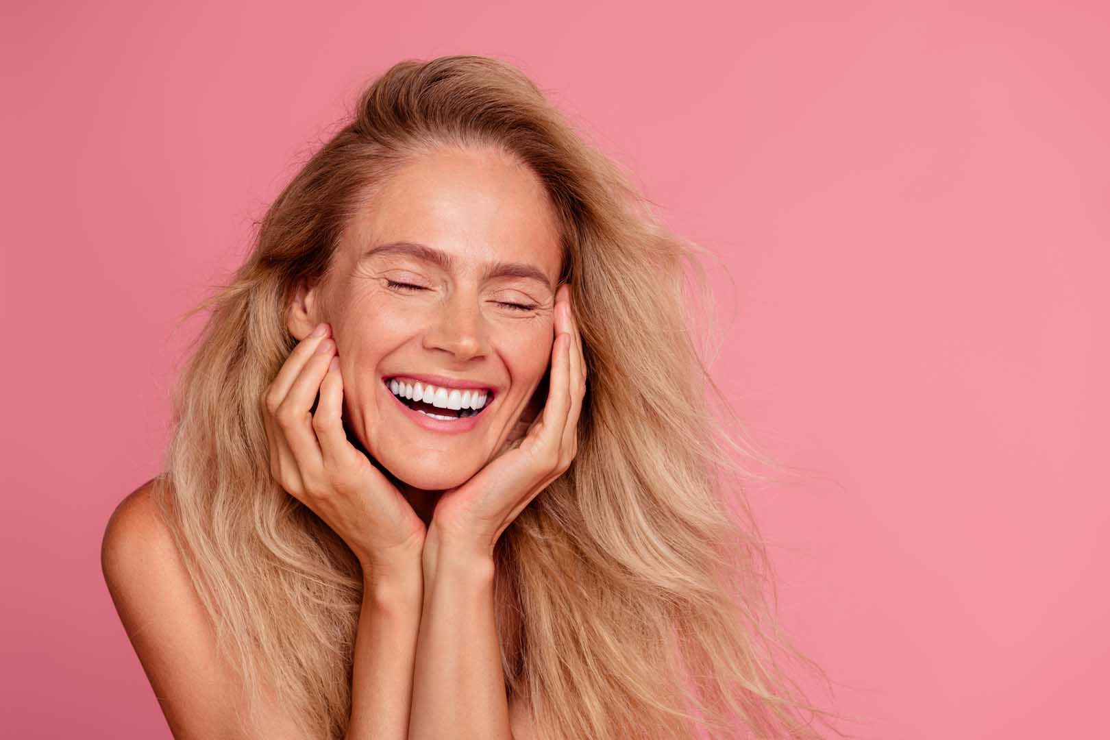 Blonde-haired person smiles widely, eyes closed, hands on cheeks, against a pink background.