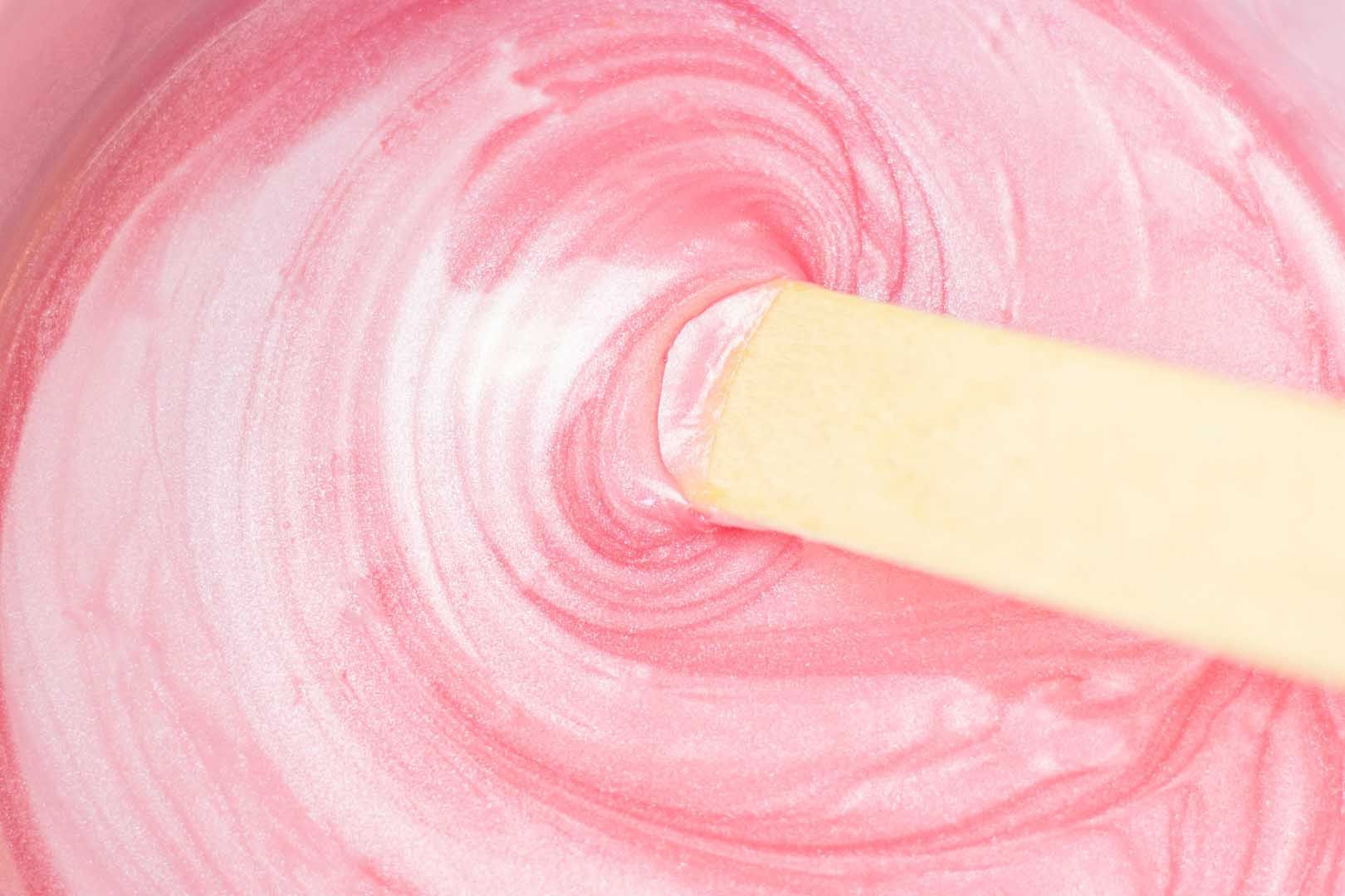 Pink, swirling wax in a container, stirred by a wooden applicator.