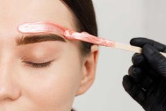 Eyebrow waxing with pink wax. Hand in black glove applies wax with a wooden stick to the brow.