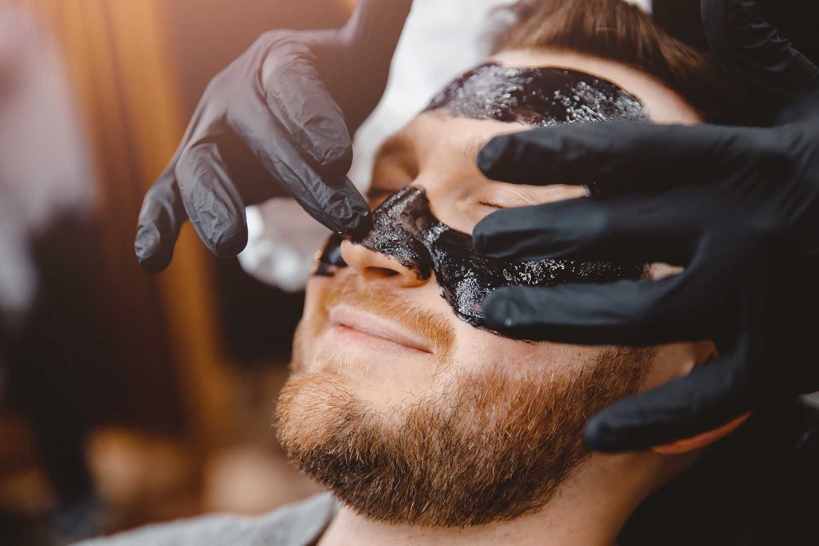 Person receiving black facial mask application; gloved hands, indoors.