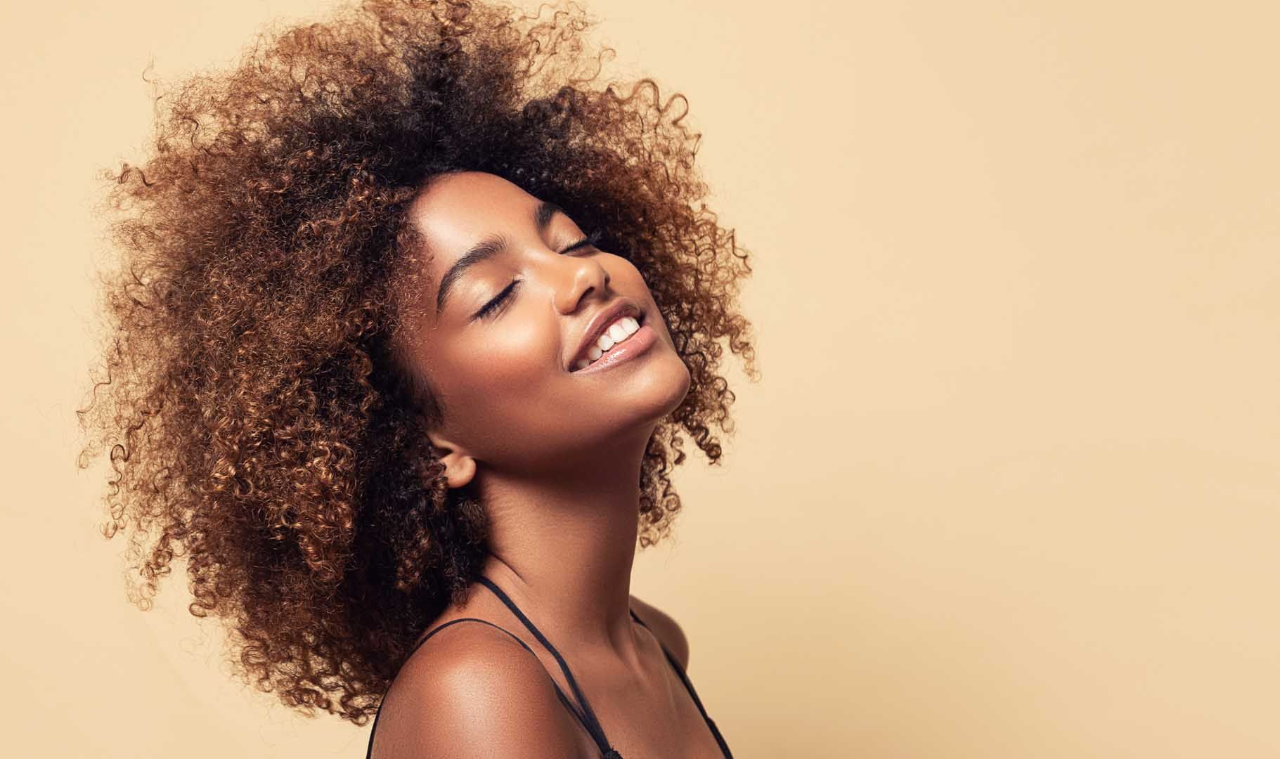 Woman with curly hair smiles, eyes closed, against a beige background.