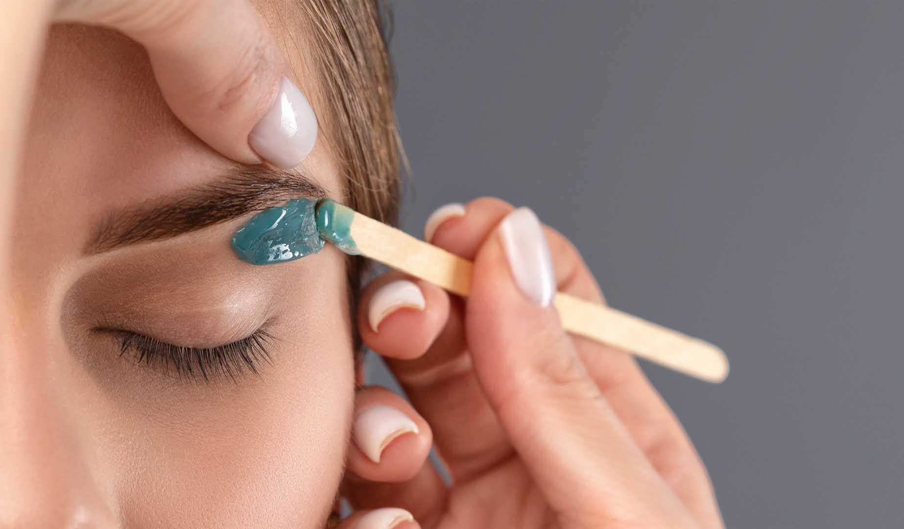 Eyebrow waxing: a person's eyebrow is coated with blue wax by a wooden stick. The person's eyes are closed.