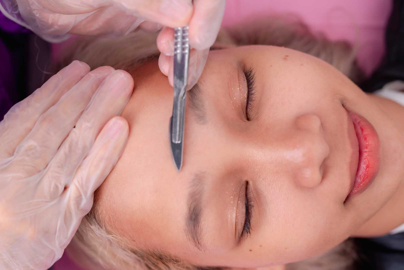 Person receiving eyebrow microblading with a blade tool; hands in gloves, close up.