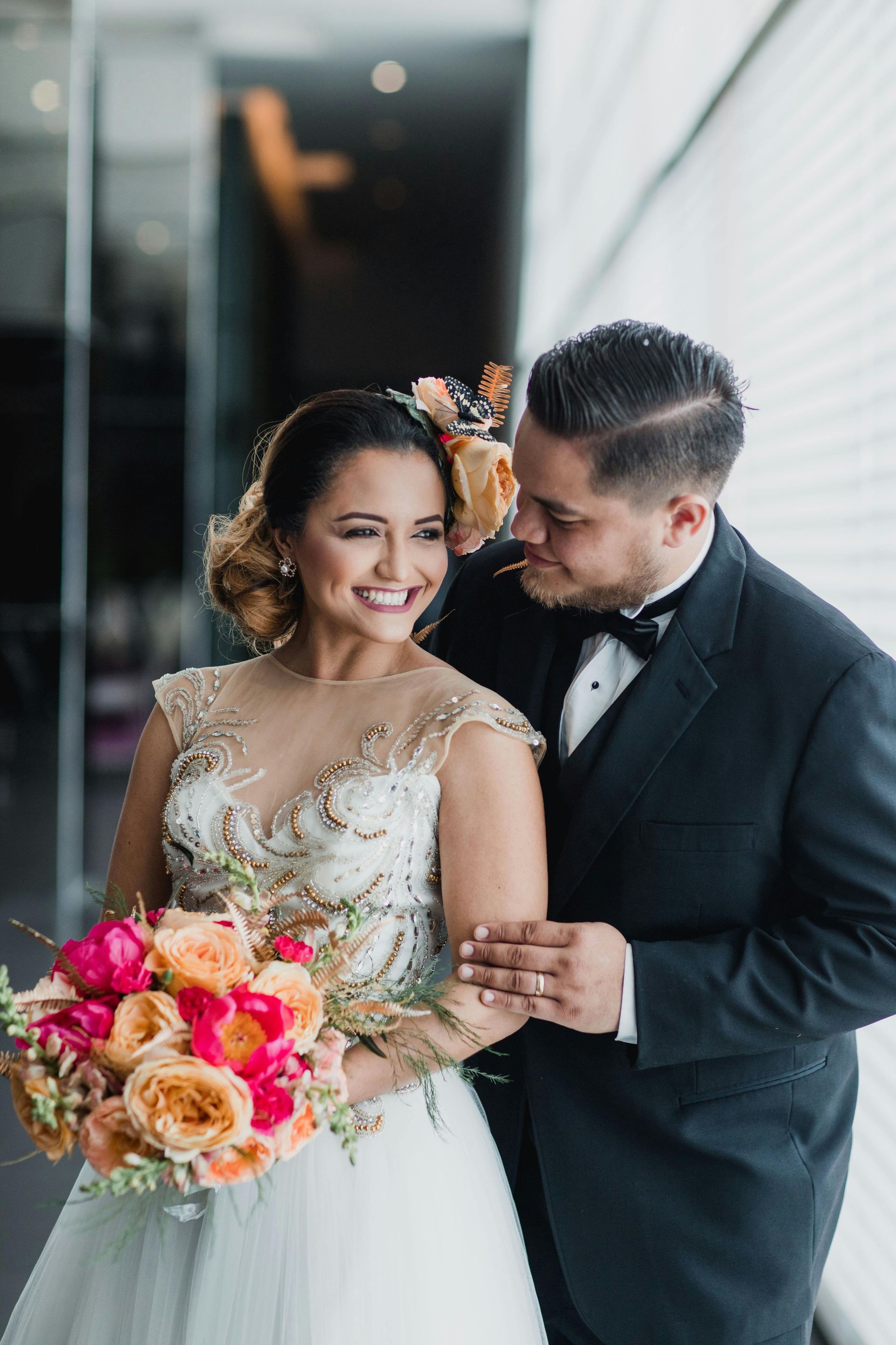 Bride and groom smile, holding a colorful bouquet, standing near a window. The groom wears a tuxedo.