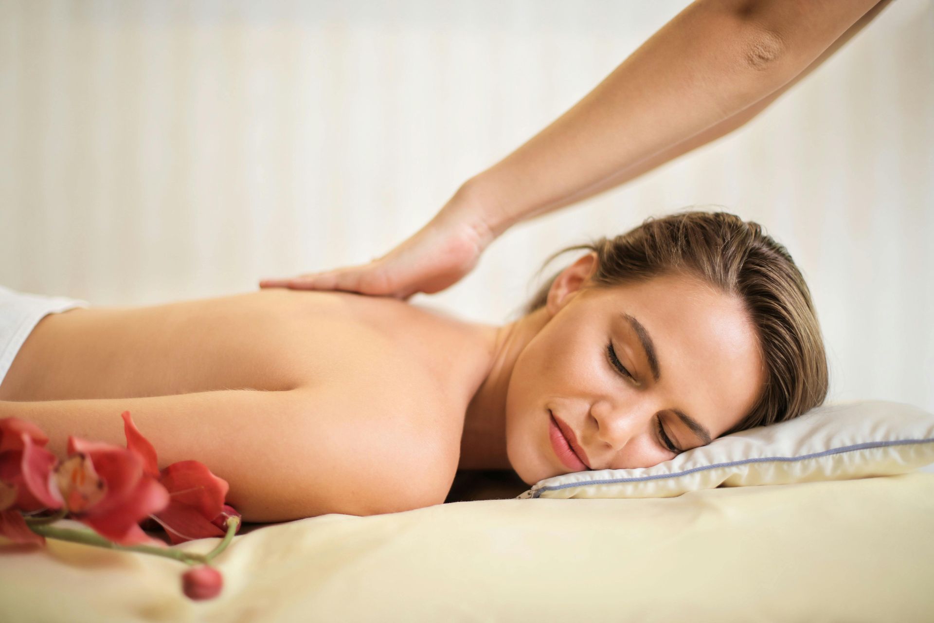 Woman receiving a back massage at a spa; hands on her back, eyes closed. Red flowers nearby.