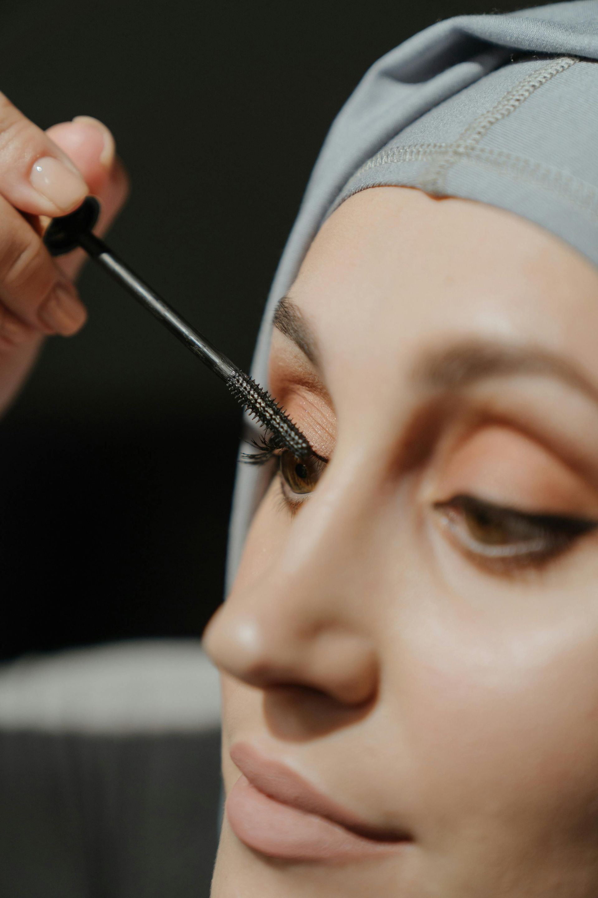 Person wearing headscarf applying mascara to eyelashes.