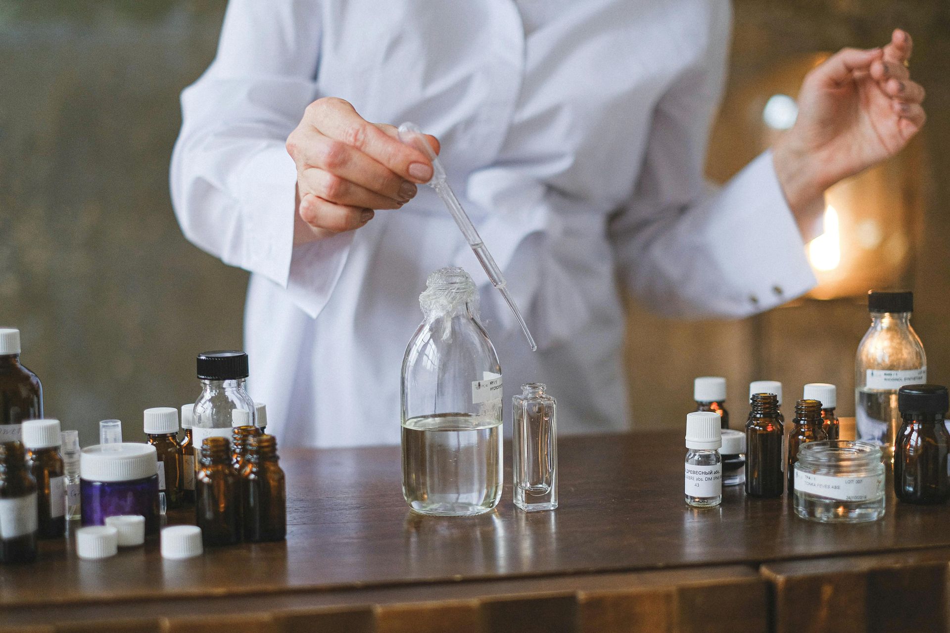 A person in a white lab coat uses a dropper to fill a bottle with liquid; various small bottles are on the table.