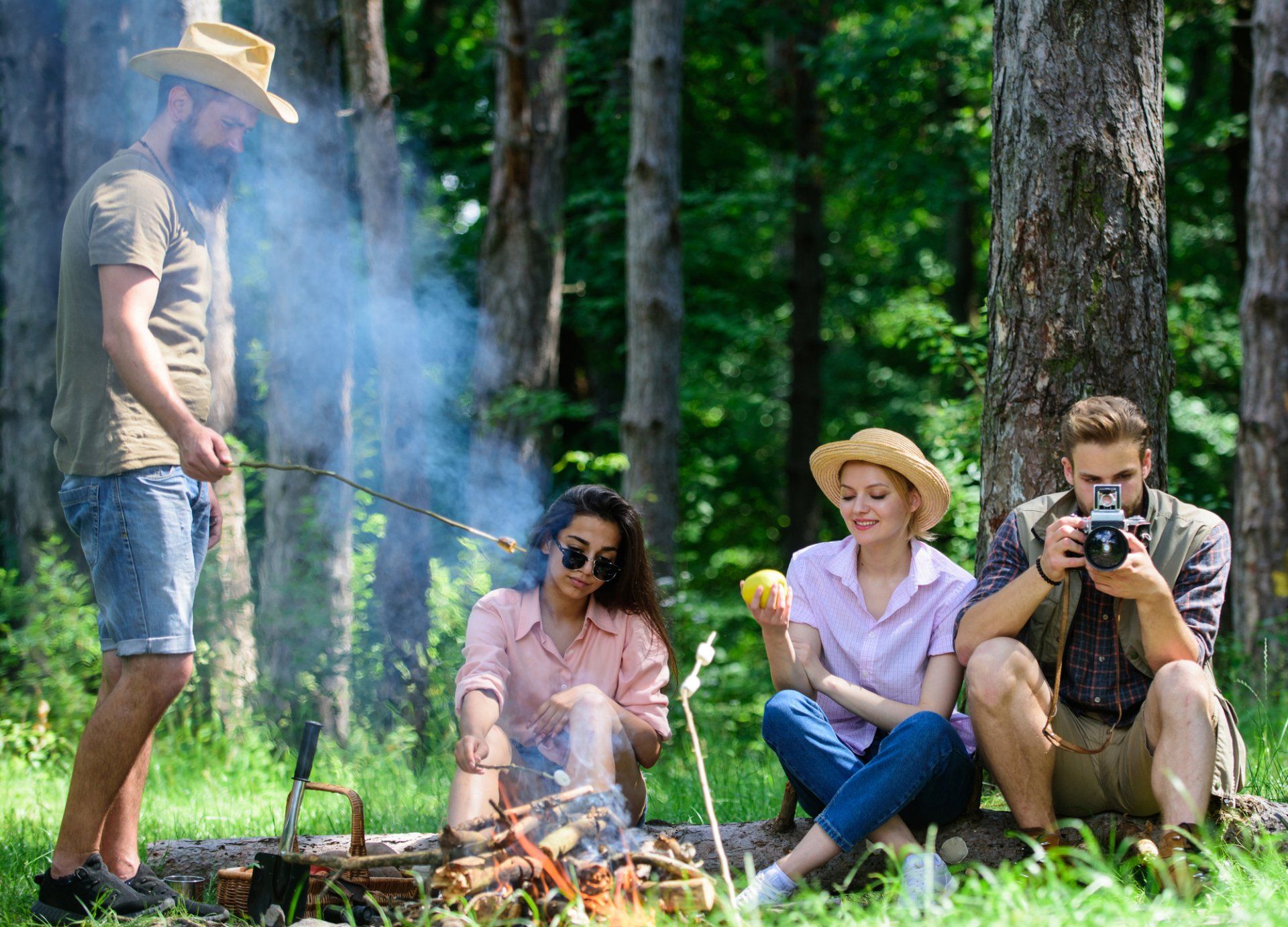 Group of friends camping in a forest, with a campfire, taking pictures, and enjoying snacks.
