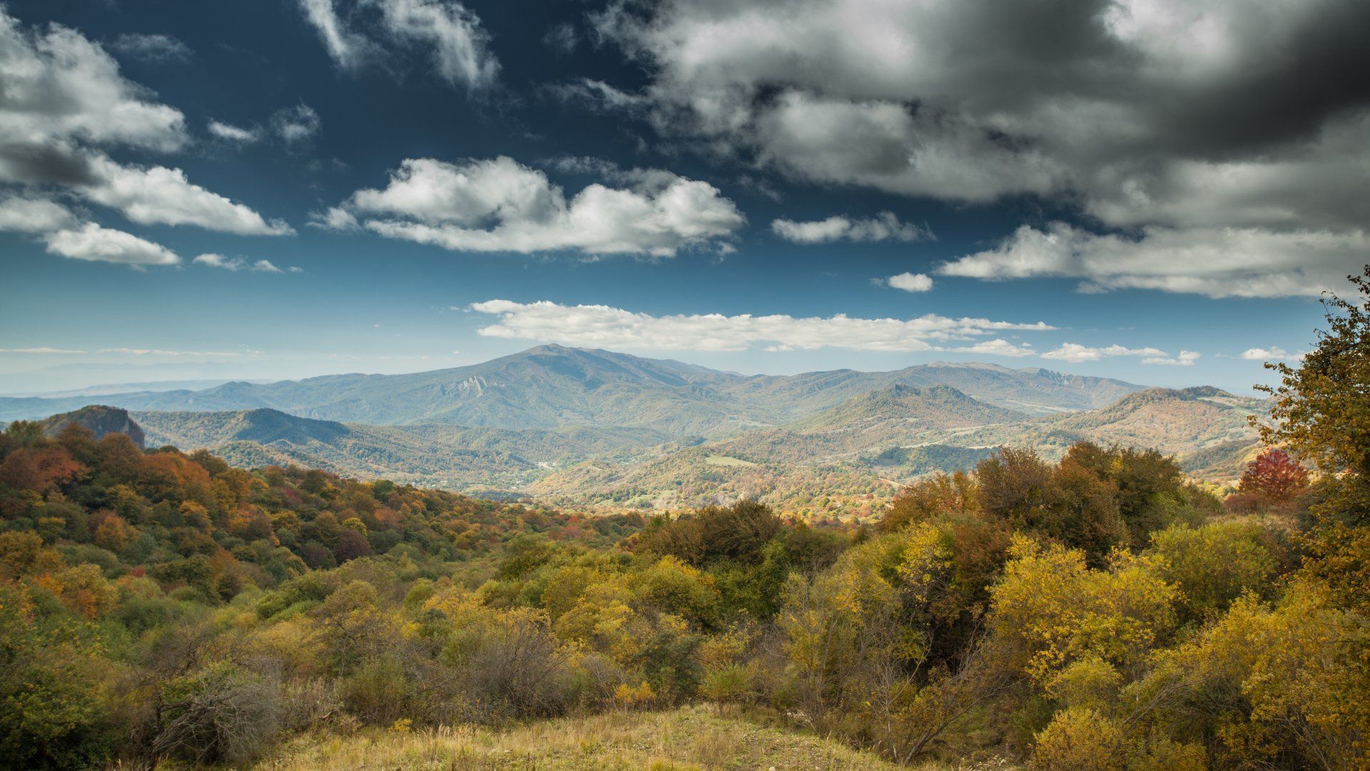 Autumn mountain landscape with trees in fall colors under a partly cloudy sky.