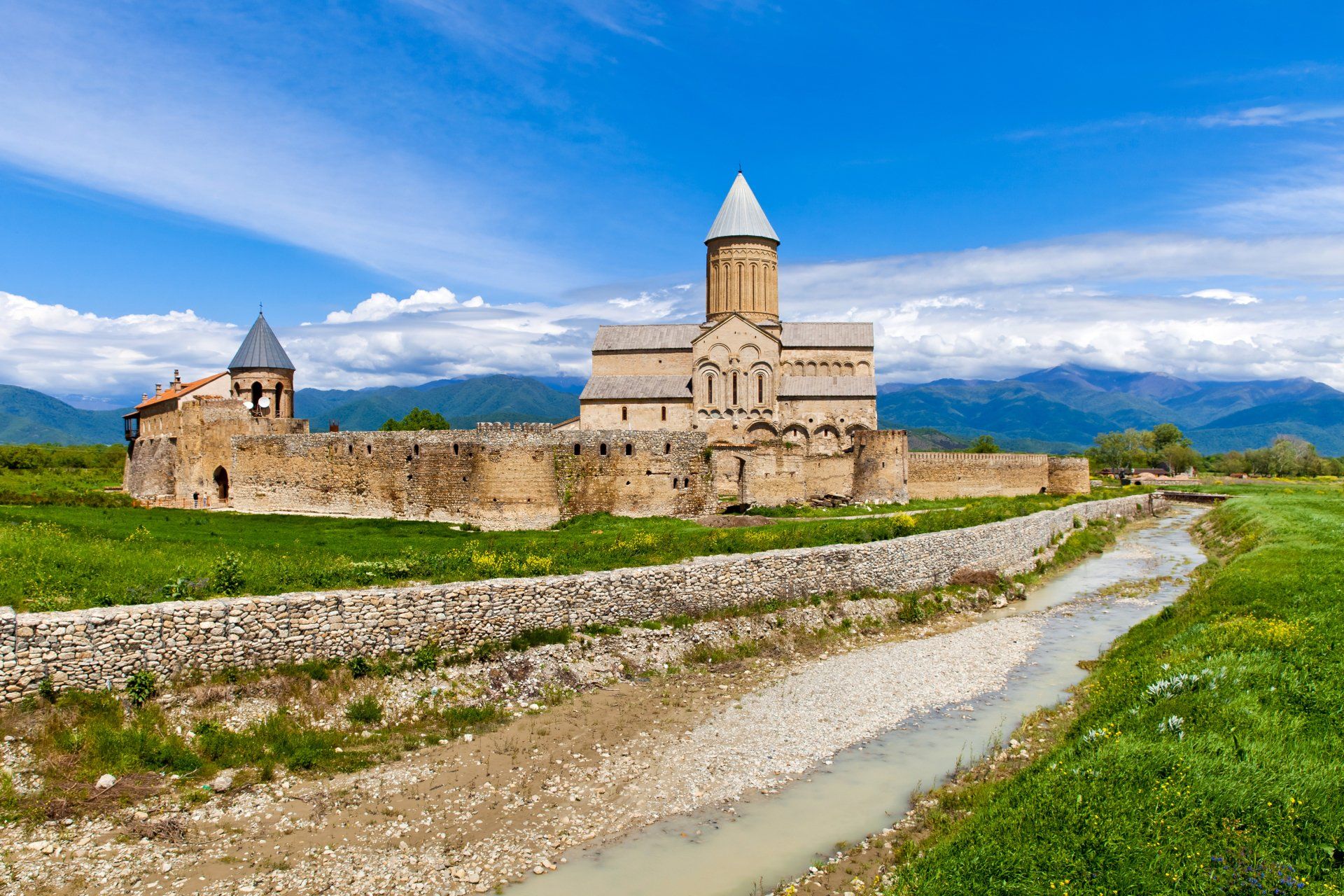 Medieval Alaverdi Monastery in Georgia, stone walls and buildings, with a small river and mountains in the background.