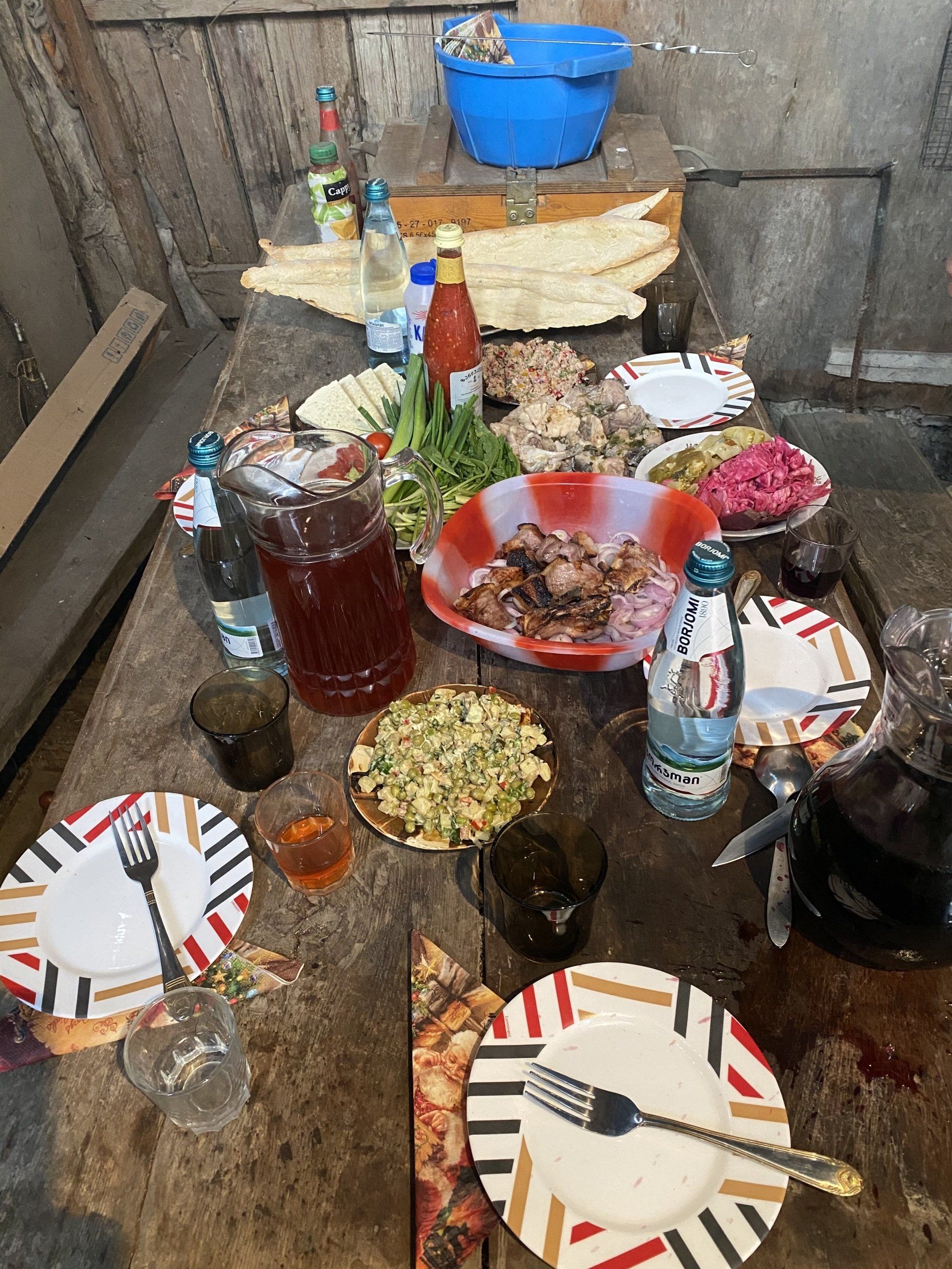 Wooden table set with food, drinks, plates, and utensils.  Bright, colorful dishes, a jug of juice, and bottles of water.