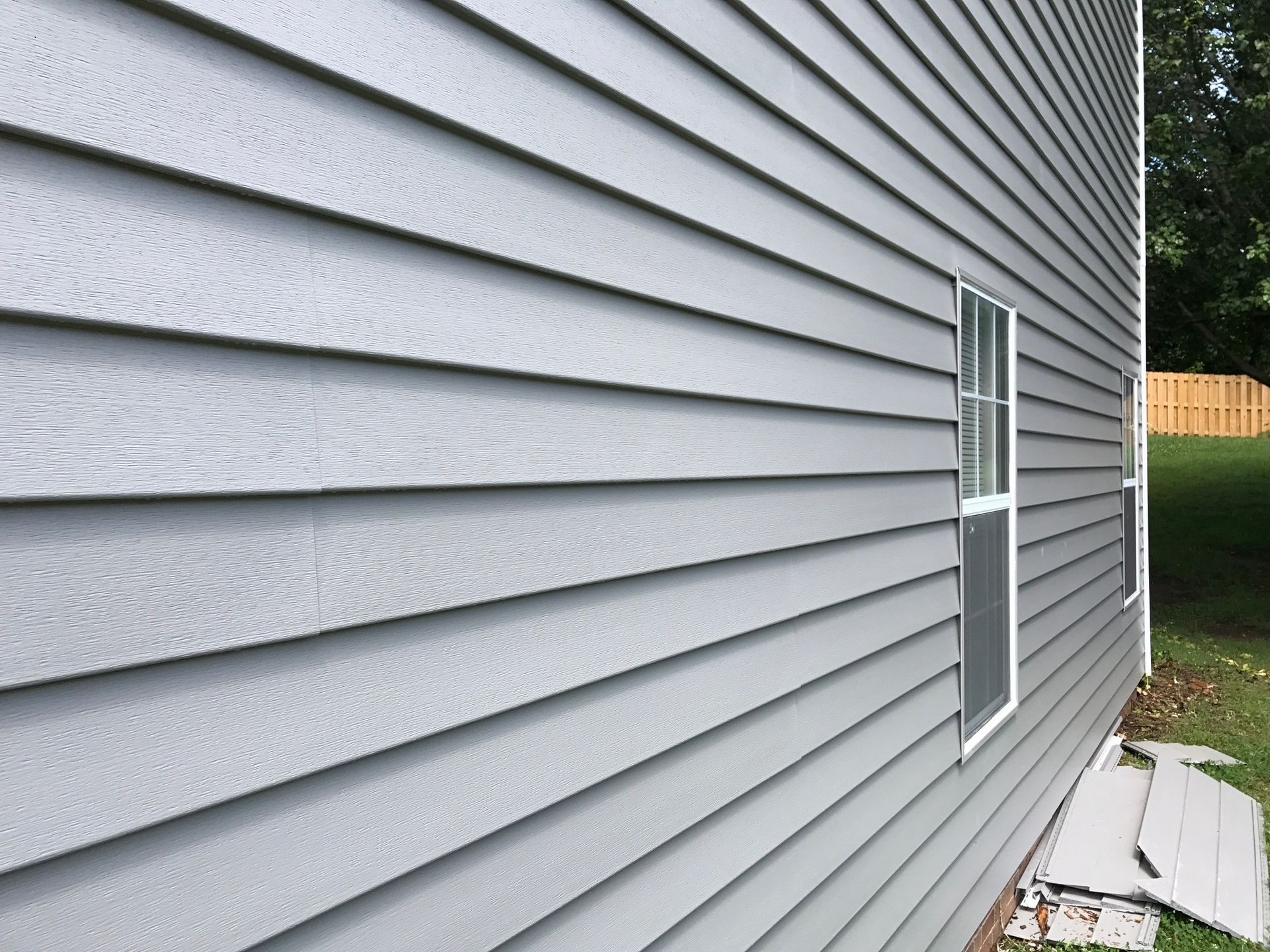 Gray siding on a house with a window, against a green yard with a fence.