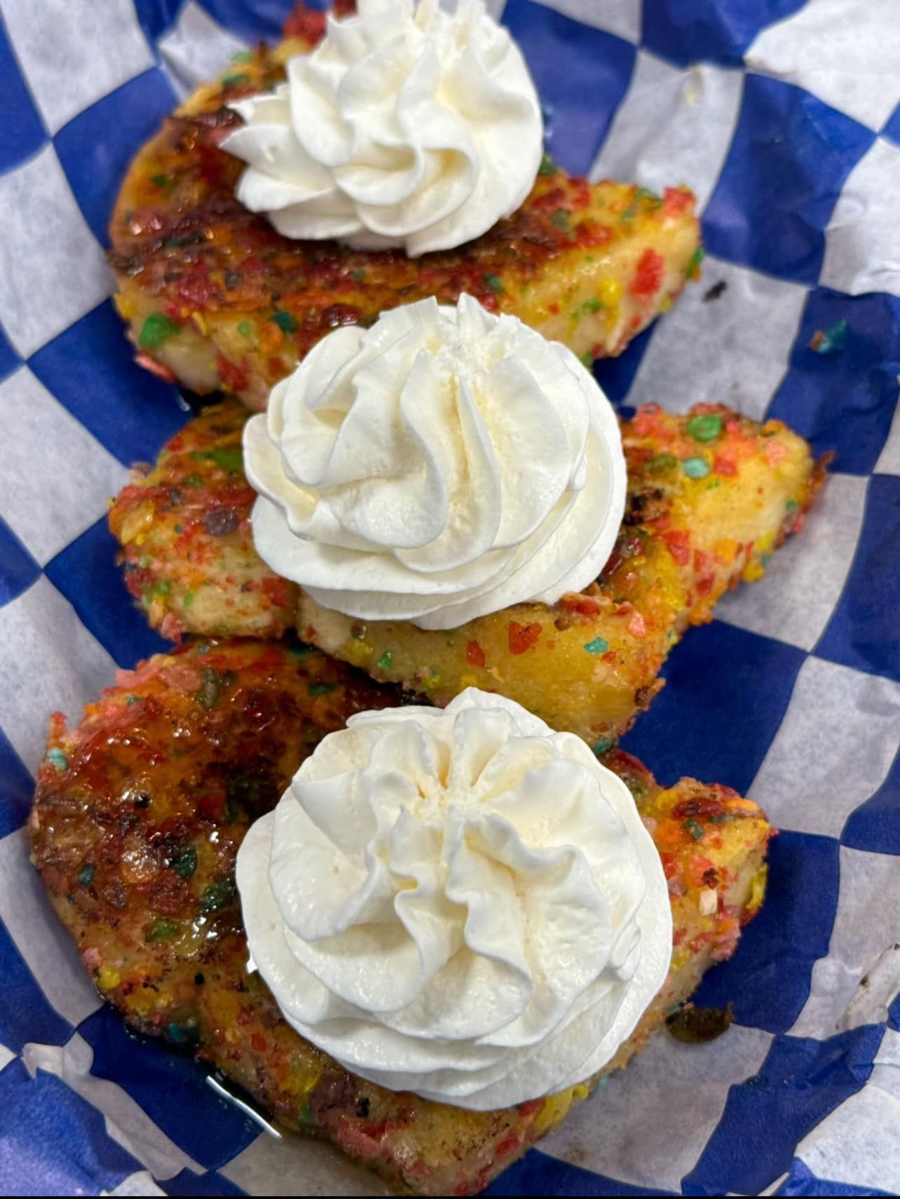 Three pieces of fried dough covered in sprinkles, topped with whipped cream, in a blue checkered paper tray.