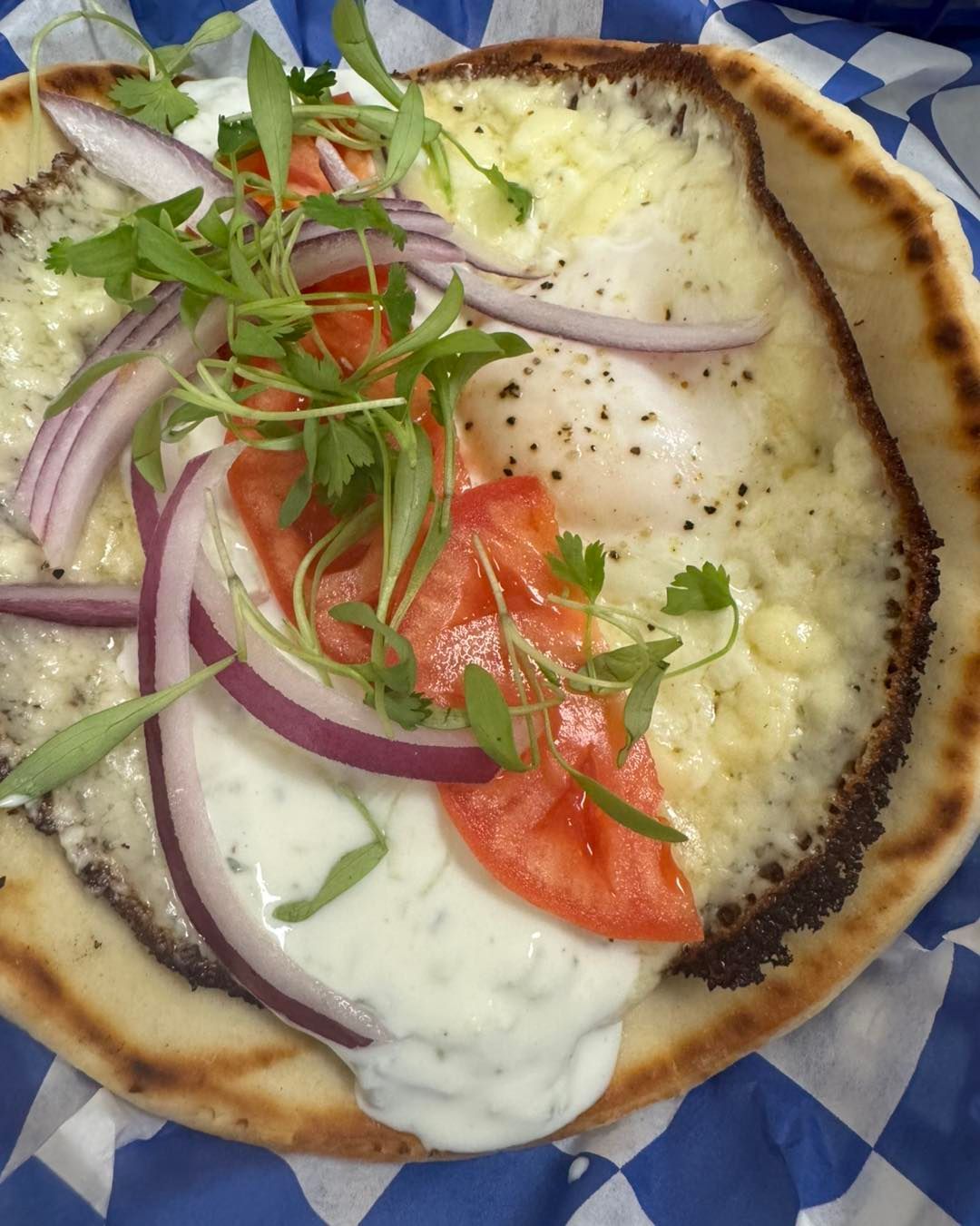 Pita bread topped with melted cheese, tomato slices, red onion, microgreens, and sauce.