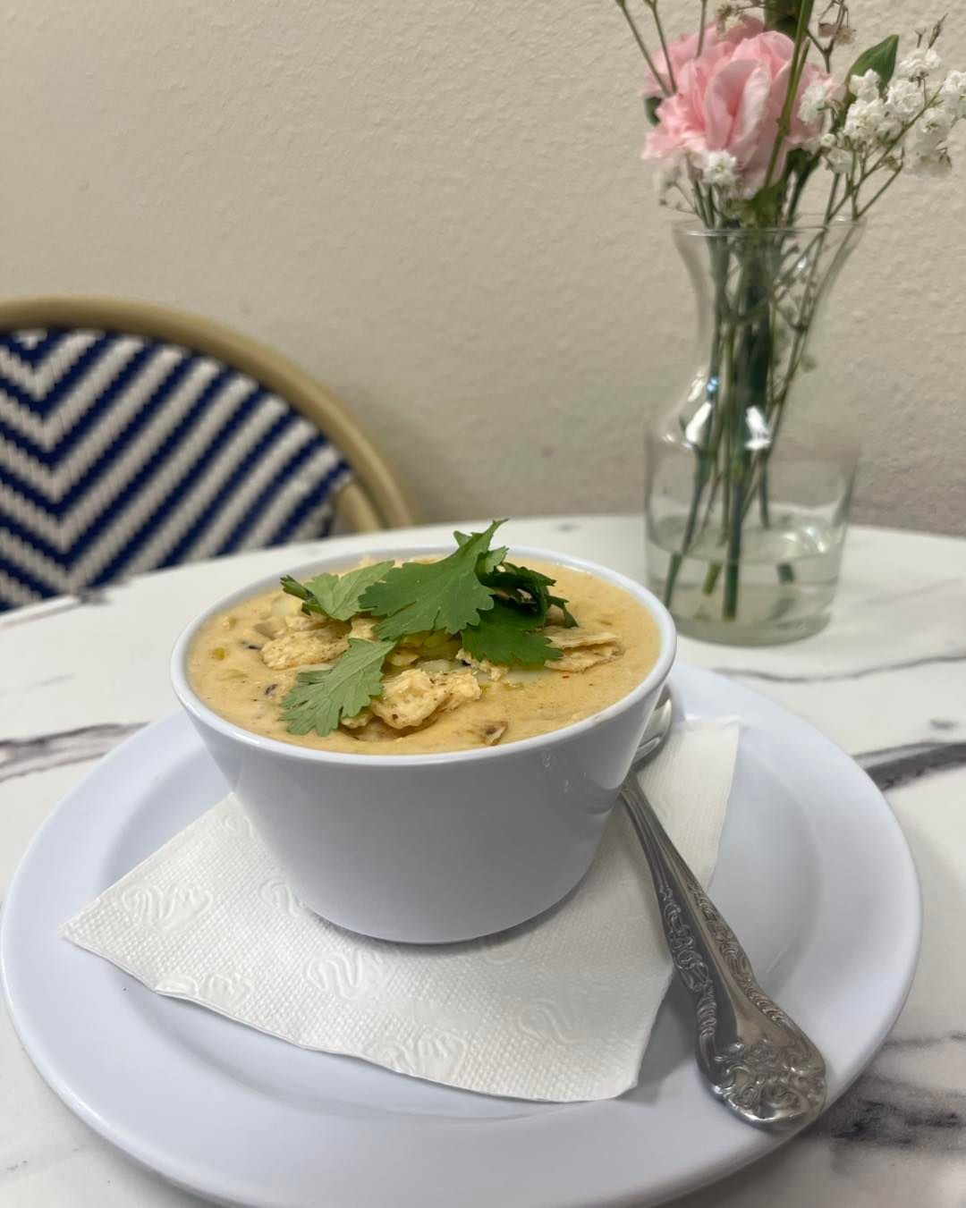 A bowl of soup with cilantro garnish on a plate, next to a vase of pink flowers on a table.