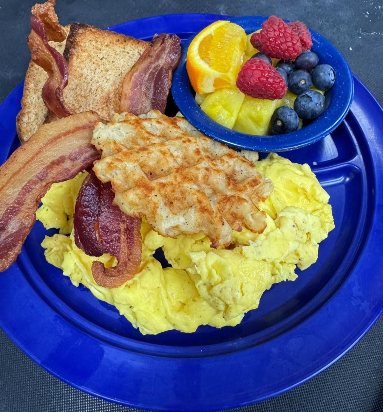 Breakfast plate with scrambled eggs, bacon, hash browns, toast, and fruit salad.