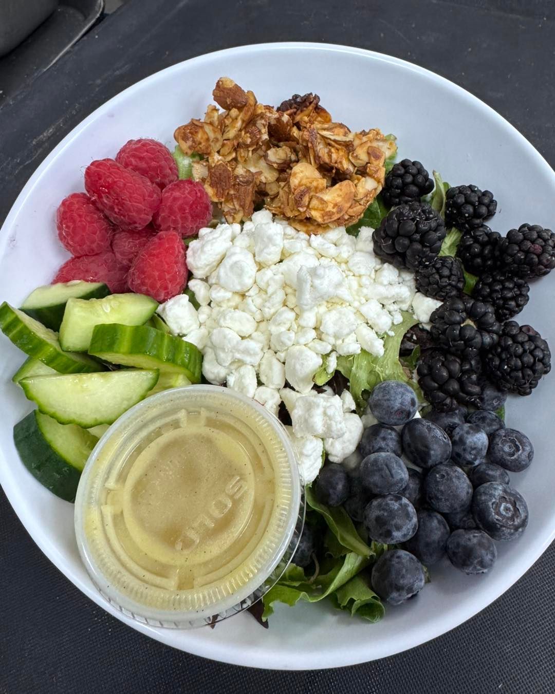Salad bowl with raspberries, blackberries, blueberries, granola, cucumber, goat cheese, and dressing.