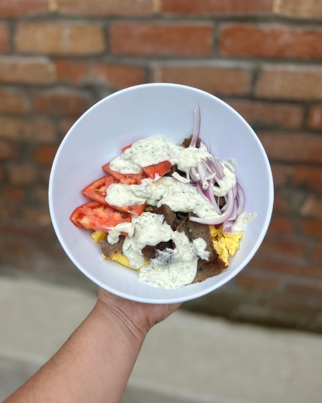 A bowl of food held by a hand, filled with tomatoes, onions, meat, and sauce against a brick wall.