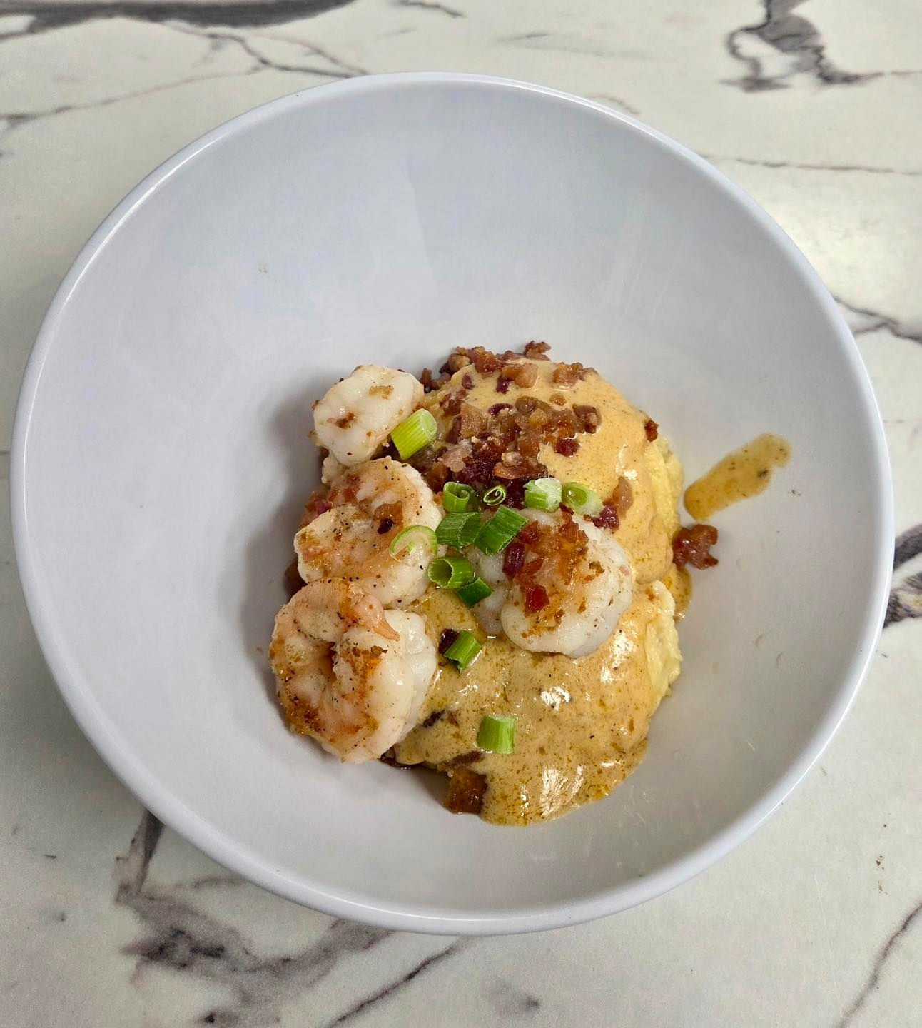 Shrimp and grits in a white bowl, topped with green onions and bacon crumbles, on a marble surface.