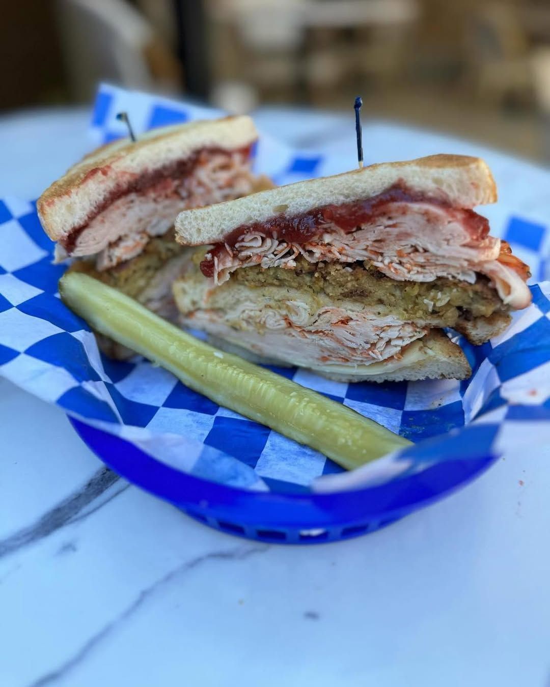 Turkey sandwich with cranberry sauce, stuffing, and pickle in a blue checkered basket.