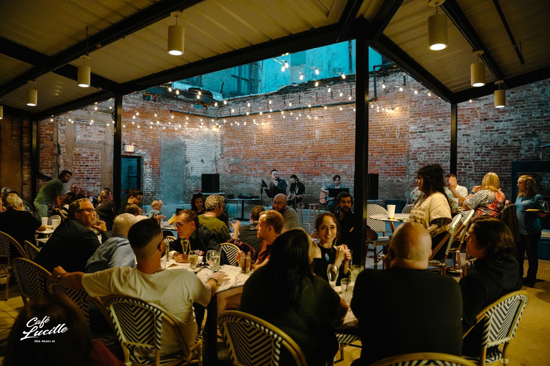 People seated at tables enjoying a live band performance in an outdoor brick-walled venue, string lights overhead.