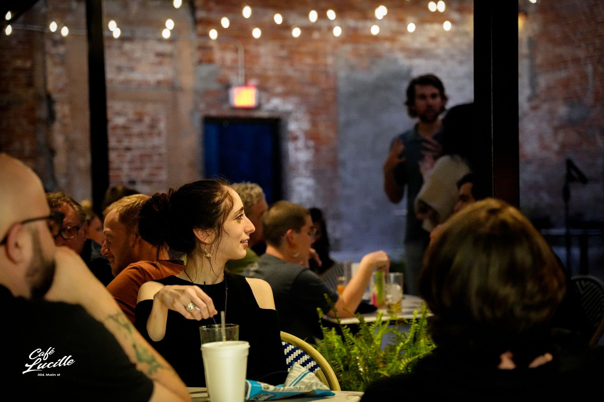 People at outdoor gathering; woman smiles, man speaks, brick wall, string lights.