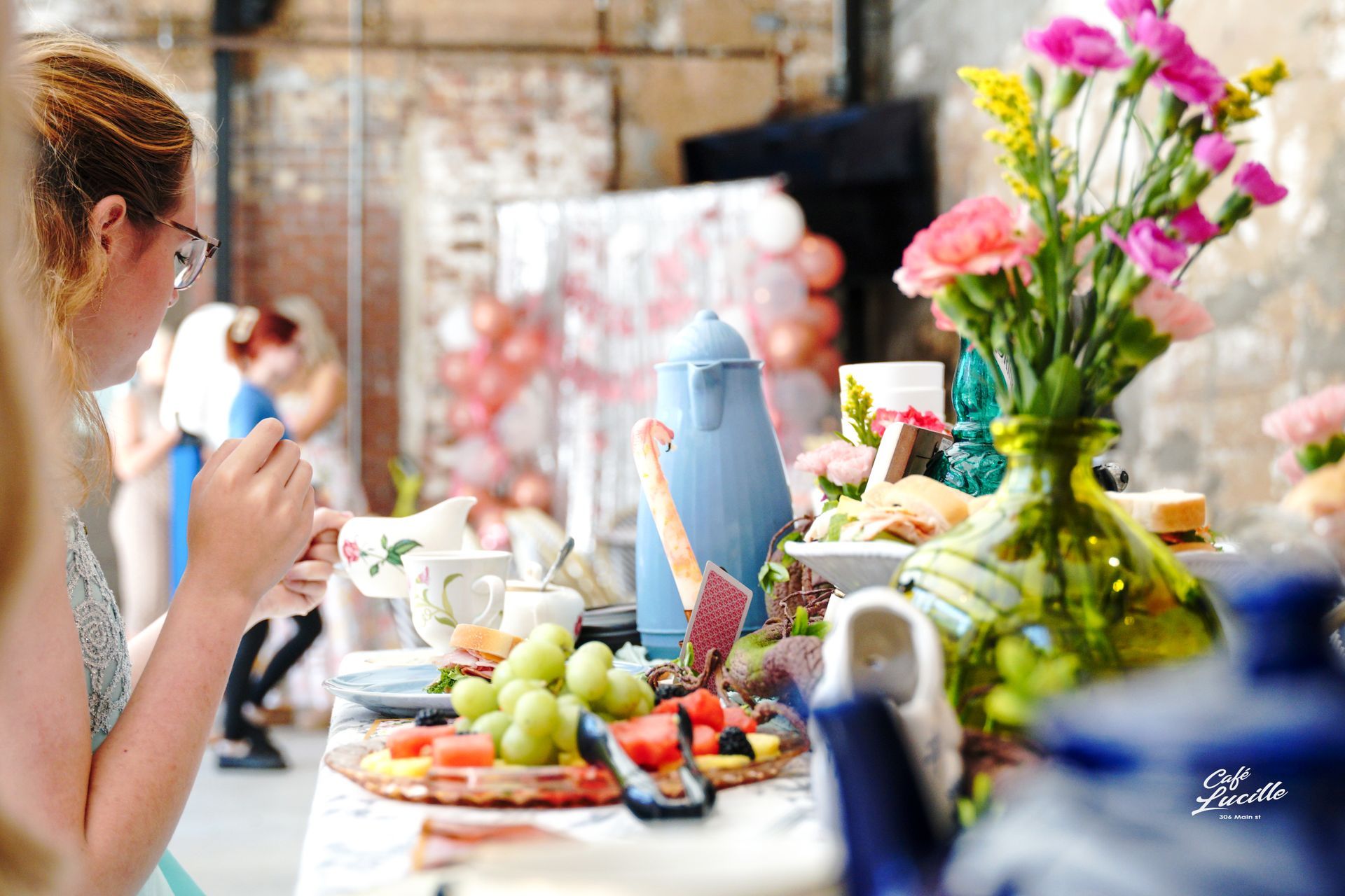 Woman at tea party, eating food from a table with fruit, flowers, and blue teapots, in a bright room.