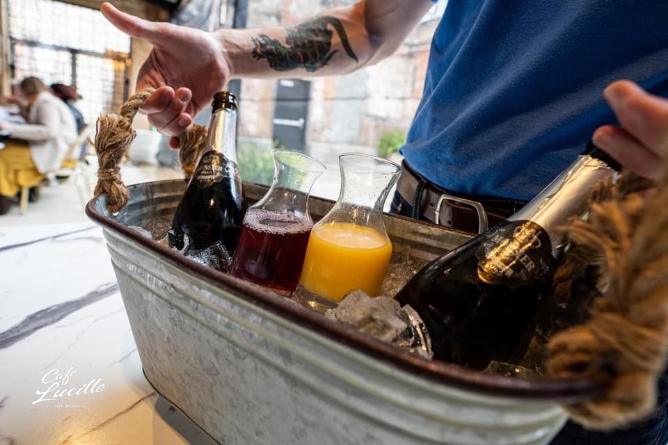 Person holding a metal tub with bottles and glasses of drinks on ice at a restaurant.