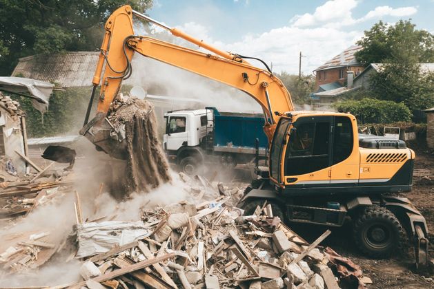 A yellow excavator is demolishing a building and loading it into a dump truck.