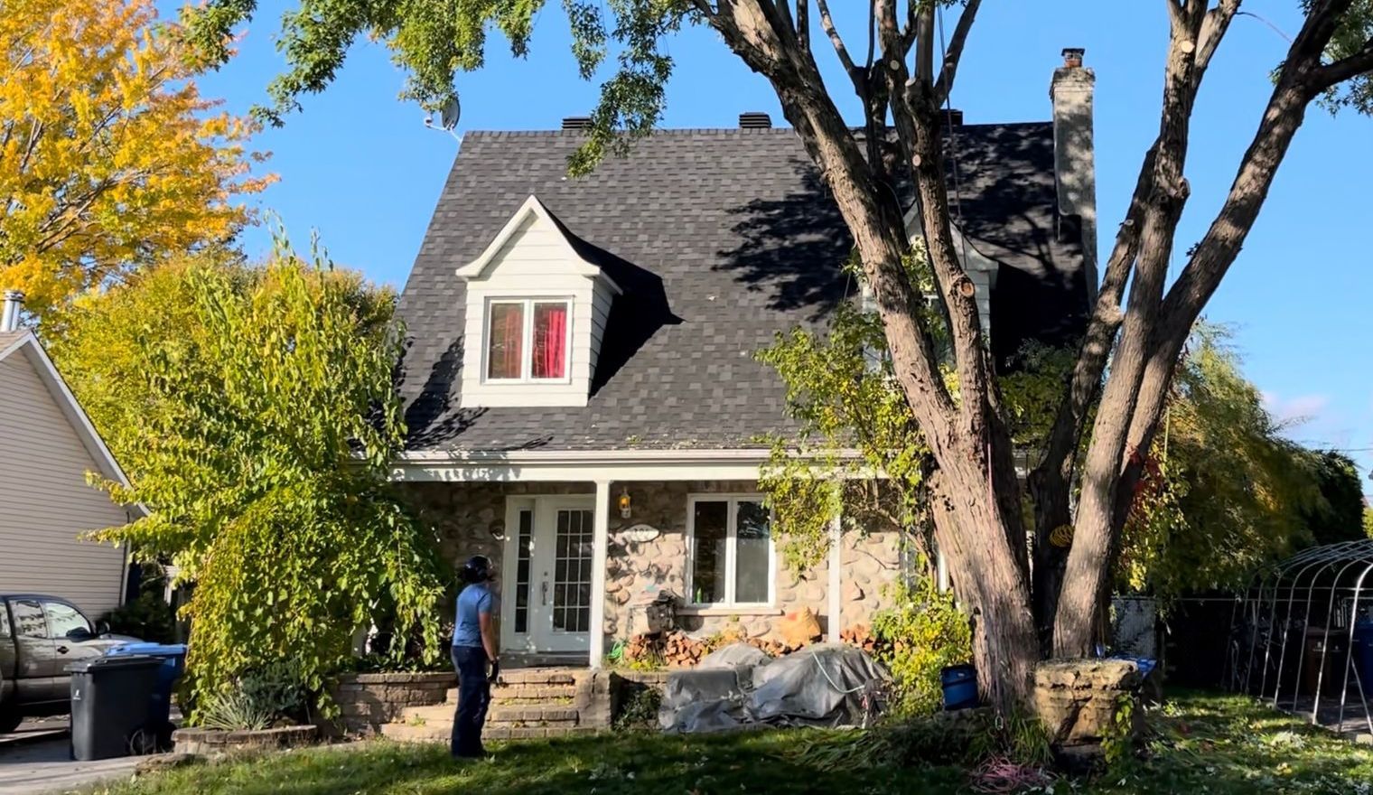 Maison en pierre au toit noir, boiseries blanches, lucarne. Une personne en bleu se tient sur des marches. Arbres, ciel bleu.