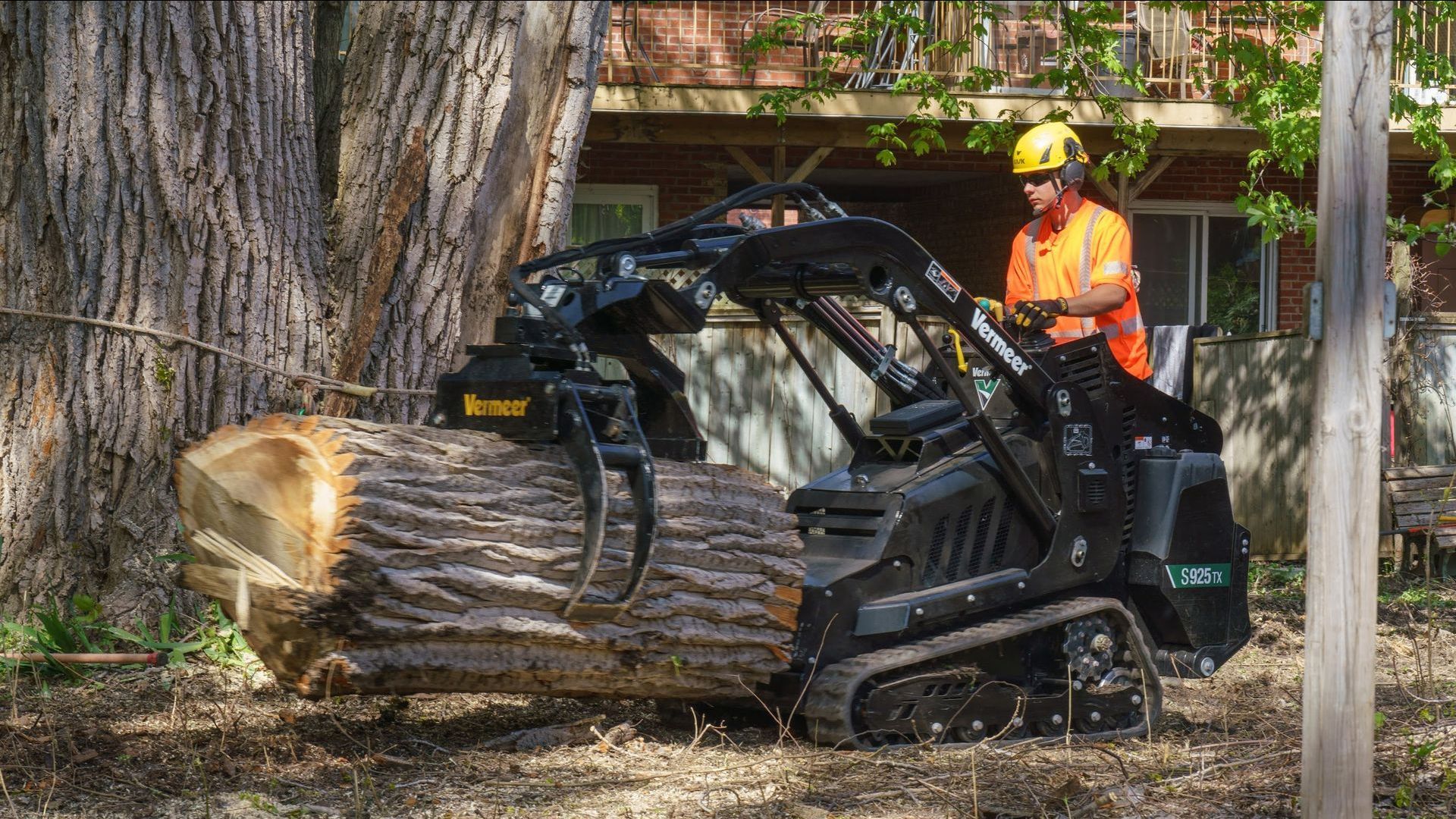 Une personne portant un gilet de sécurité orange manœuvre une petite chargeuse compacte équipée d'une pince à grumes, déplaçant un tronc d'arbre coupé.