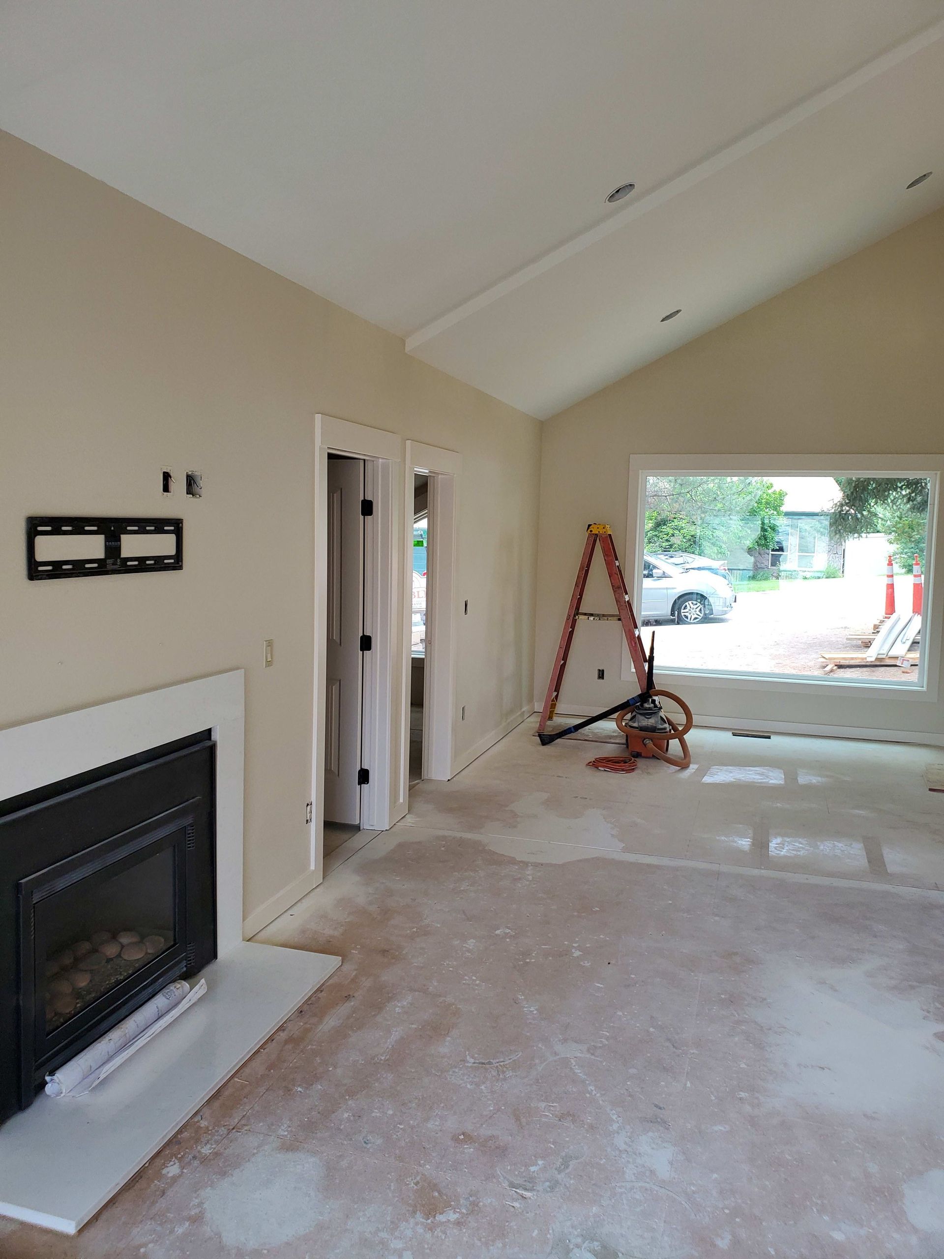 Living room renovation with a fireplace, window, and ladder. Walls are beige, floors bare.