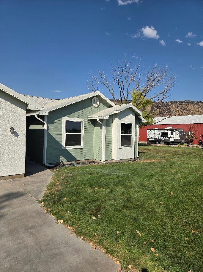 Green house with white trim, windows, and a sloped roof, on a grassy lawn, with a red building in the background.