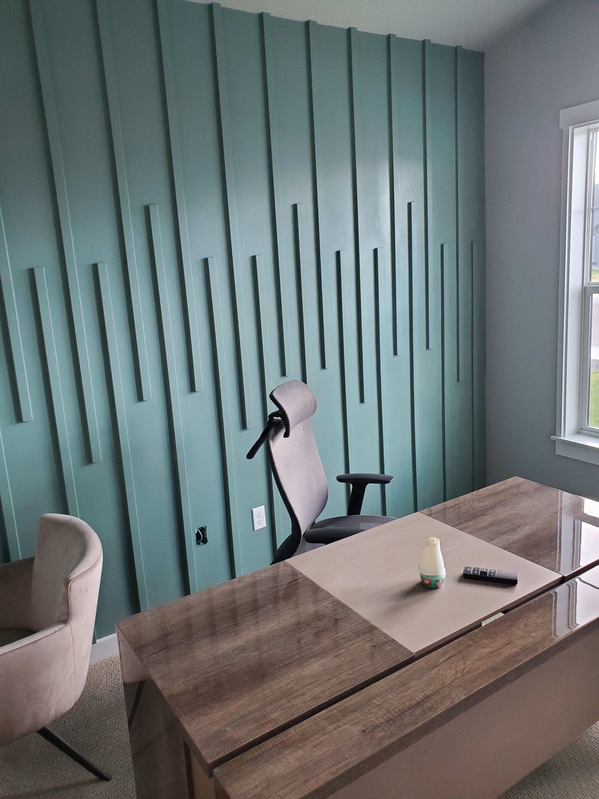 A teal accent wall with vertical wood panels behind a desk and office chair in a well-lit office space.