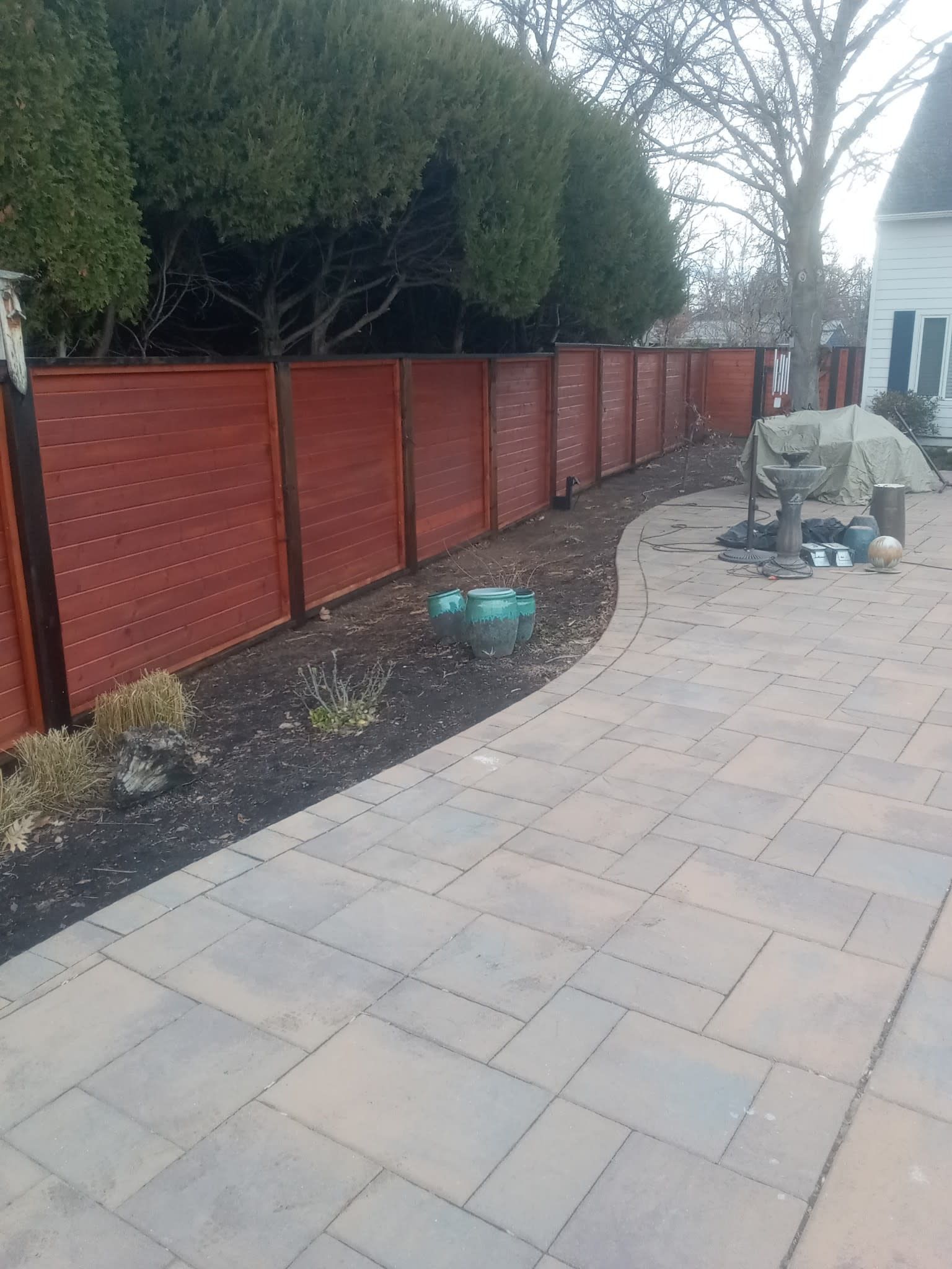 Red stained wooden fence along a patio with a dark border, and evergreen trees in the background.