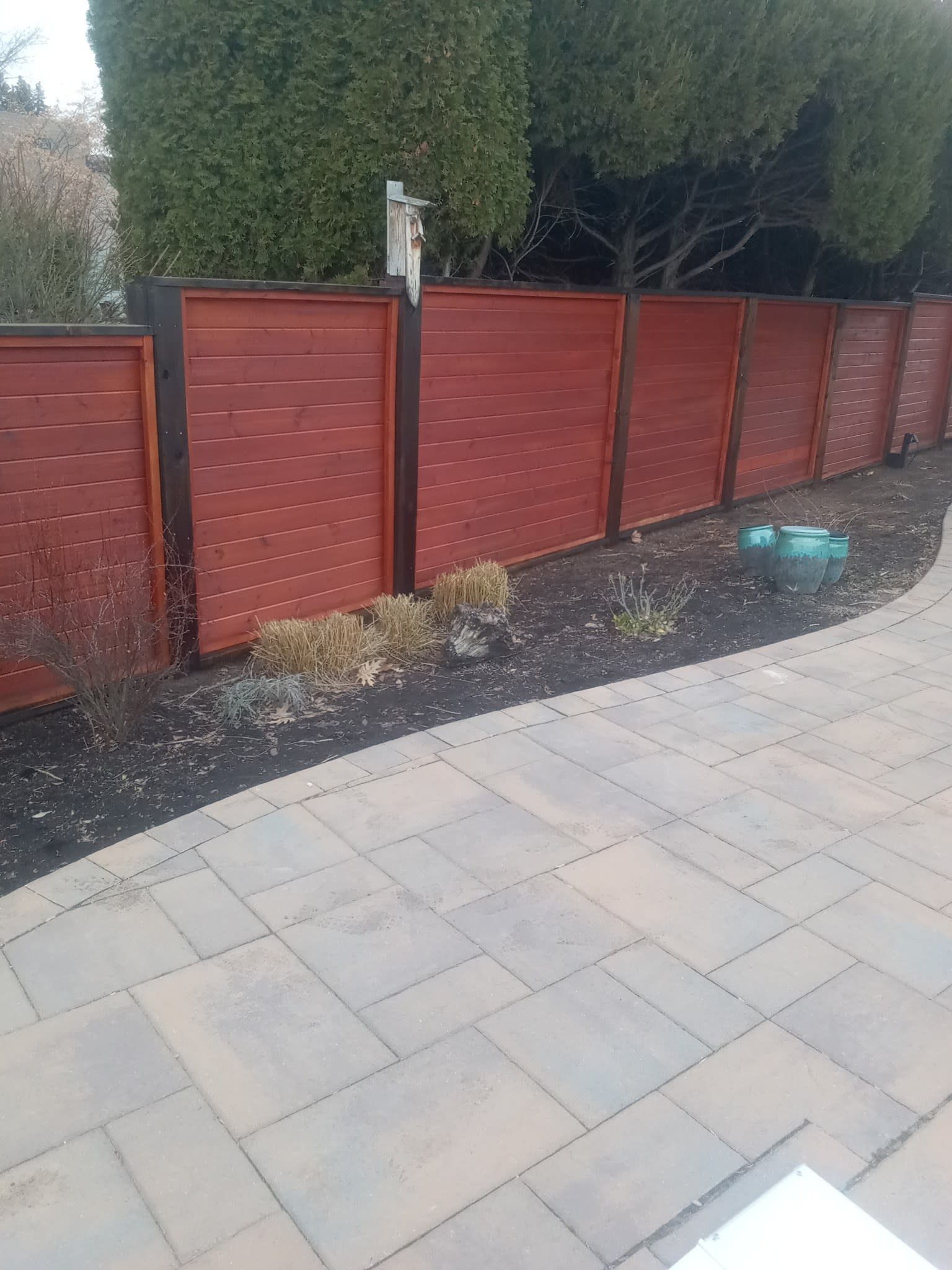 Red horizontal slat fence along a patio with pavers and a green shrub background.