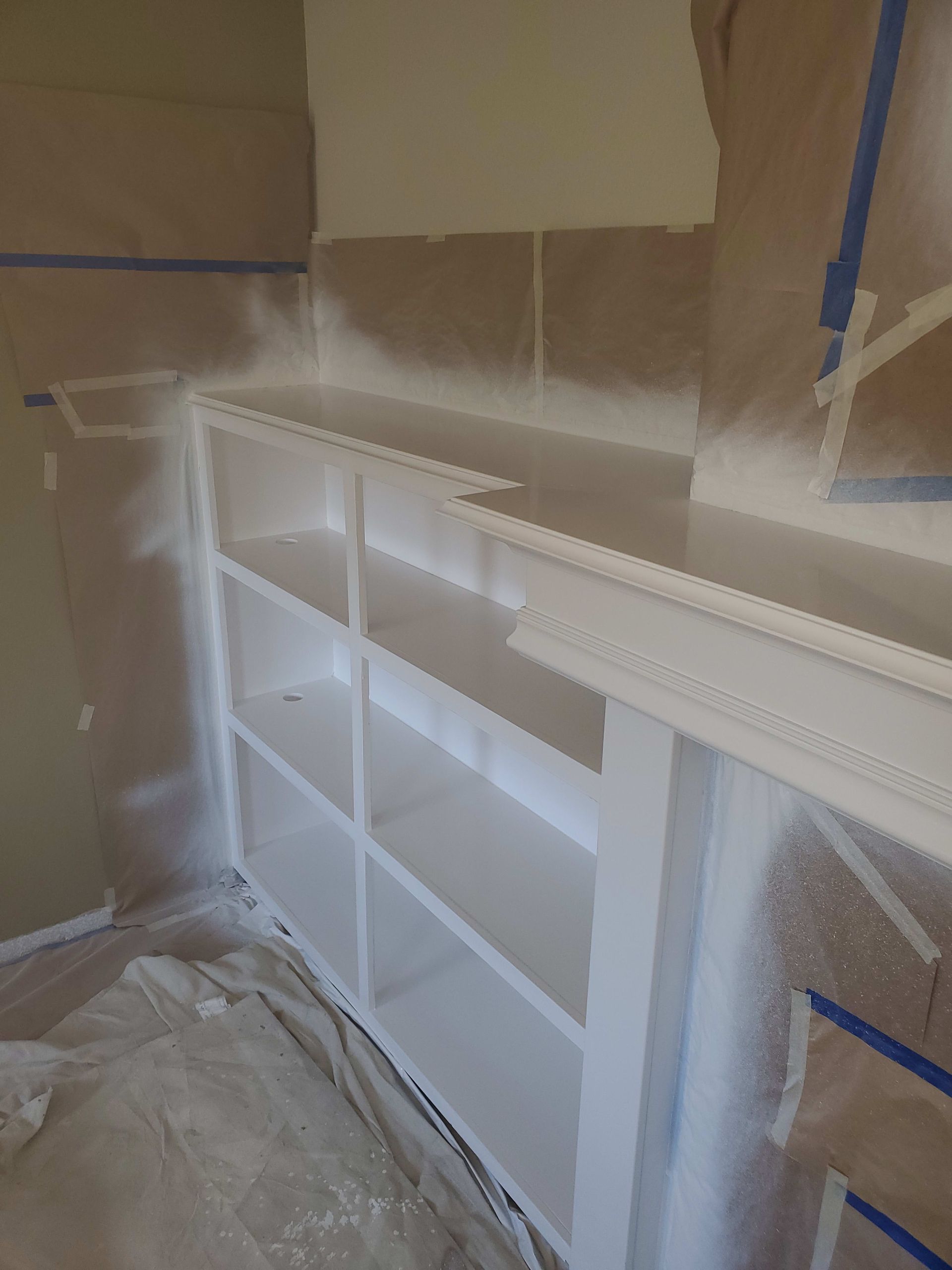 White bookcase being painted with a spray gun, walls and floor protected by paper, blue tape visible.