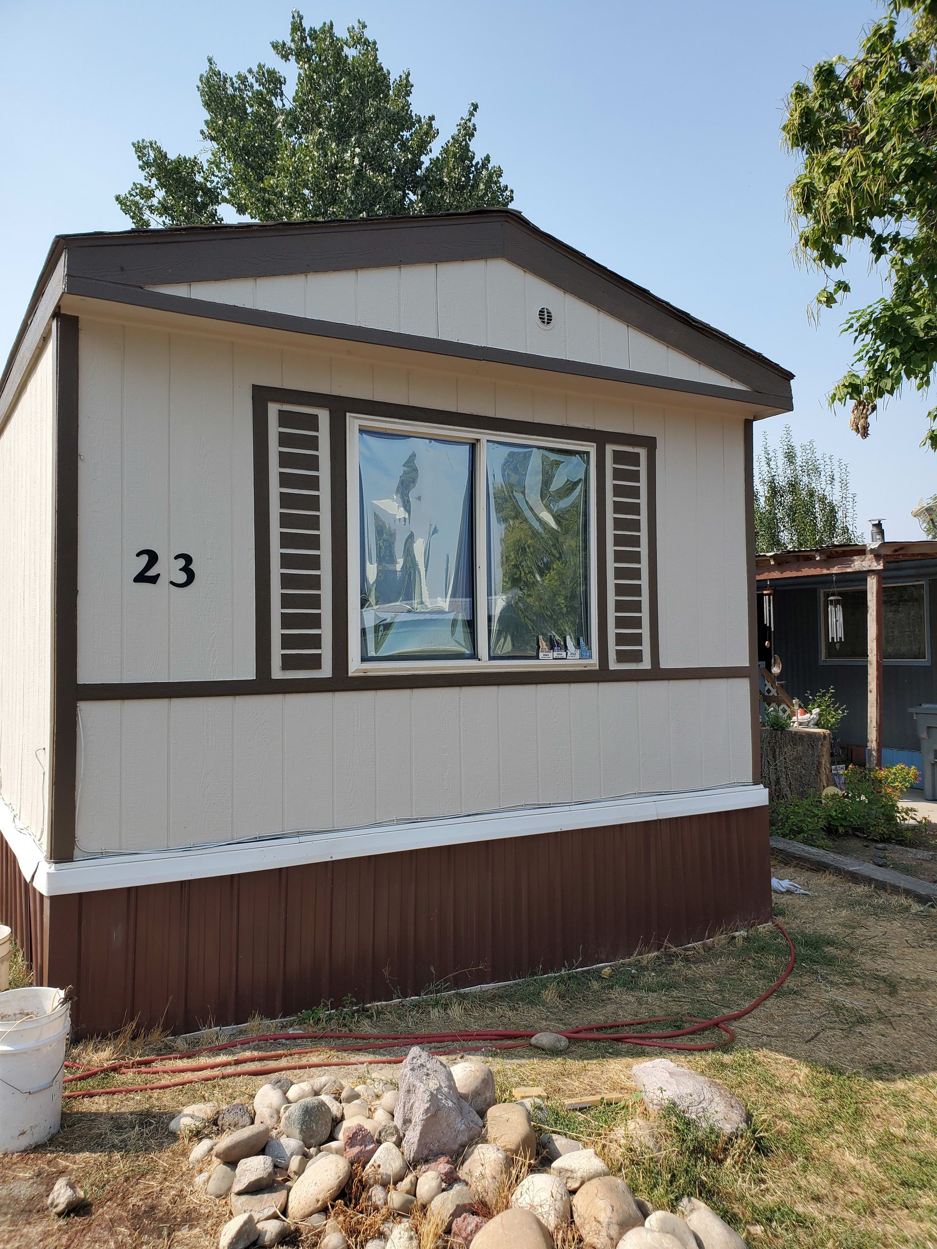 Tan and brown mobile home with number 23 on it, with window, shutters, and rocky foreground.