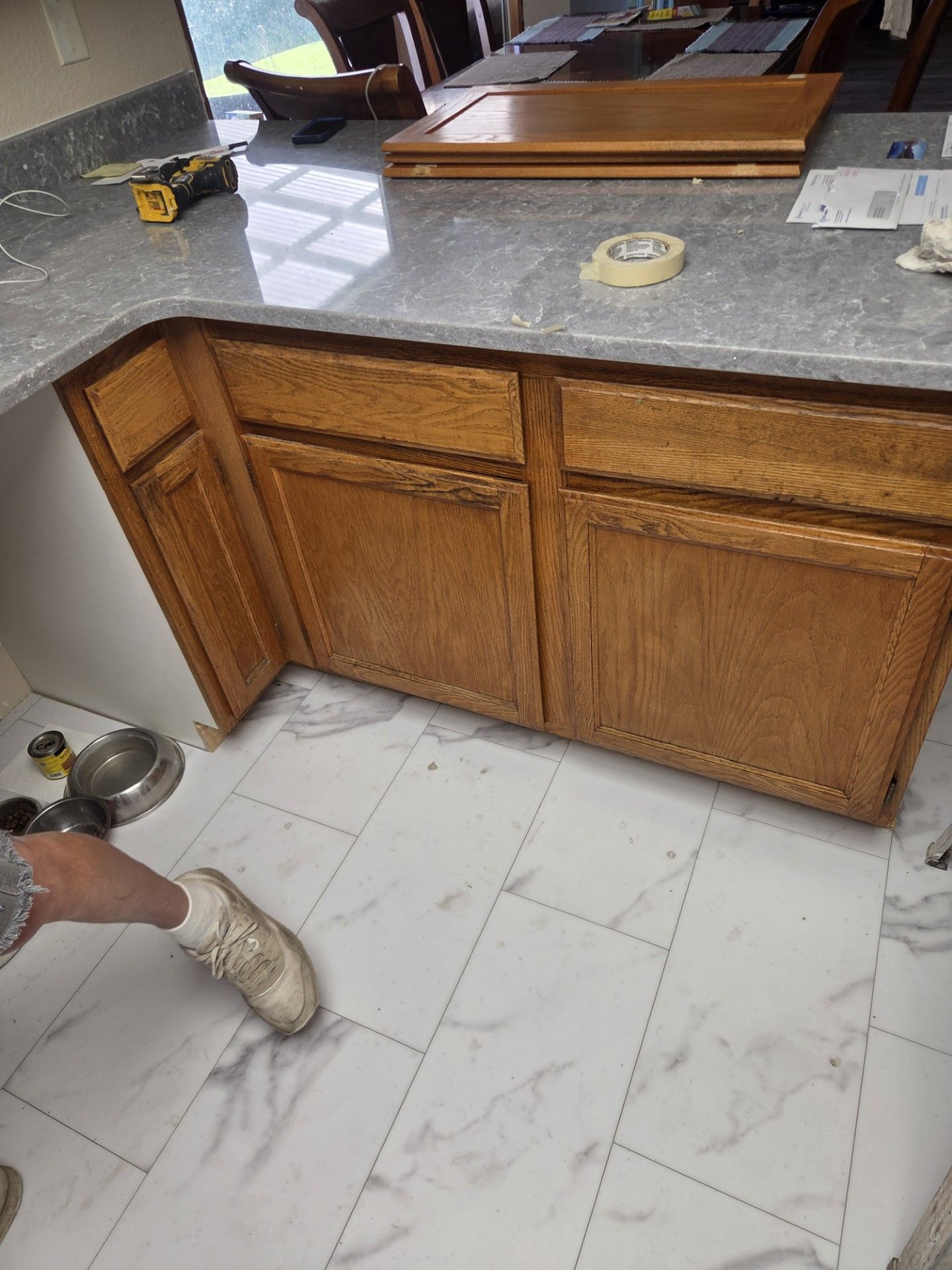 Kitchen cabinets with wood doors and a gray countertop, a person's leg and foot near new white flooring.