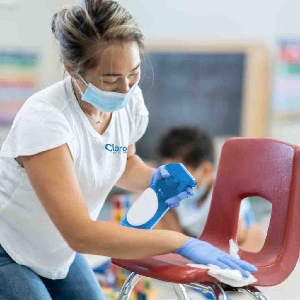 Une femme portant un masque et des gants désinfecte une chaise rouge dans une salle de classe.