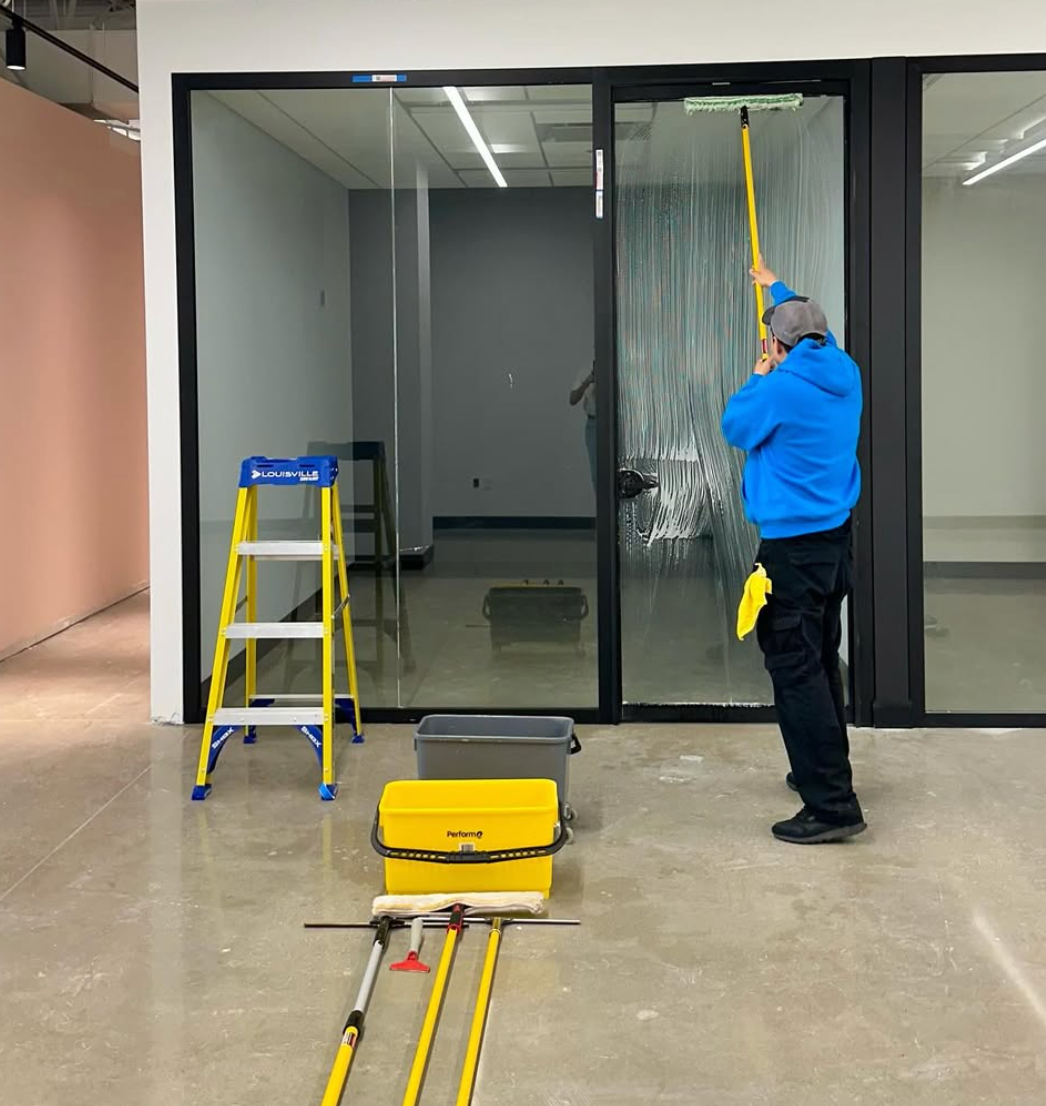 Un homme passe l'aspirateur dans un bureau moquetté avec un aspirateur dorsal. La pièce est meublée d'une table de conférence et de chaises.