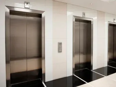 Dans un couloir, trois portes d'ascenseur en acier inoxydable sont identifiées par les lettres A, B et C. Le sol est noir et blanc.