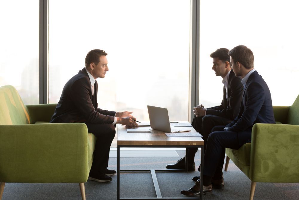 Three Men in Suits Sit at a Table, Engaged in a Discussion Near a Window — Search Partners in Melbourne, VIC