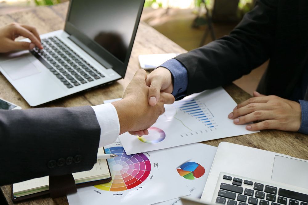 Two Men in Suits Shaking Hands Over a Table With Laptops — Search Partners in Melbourne, VIC